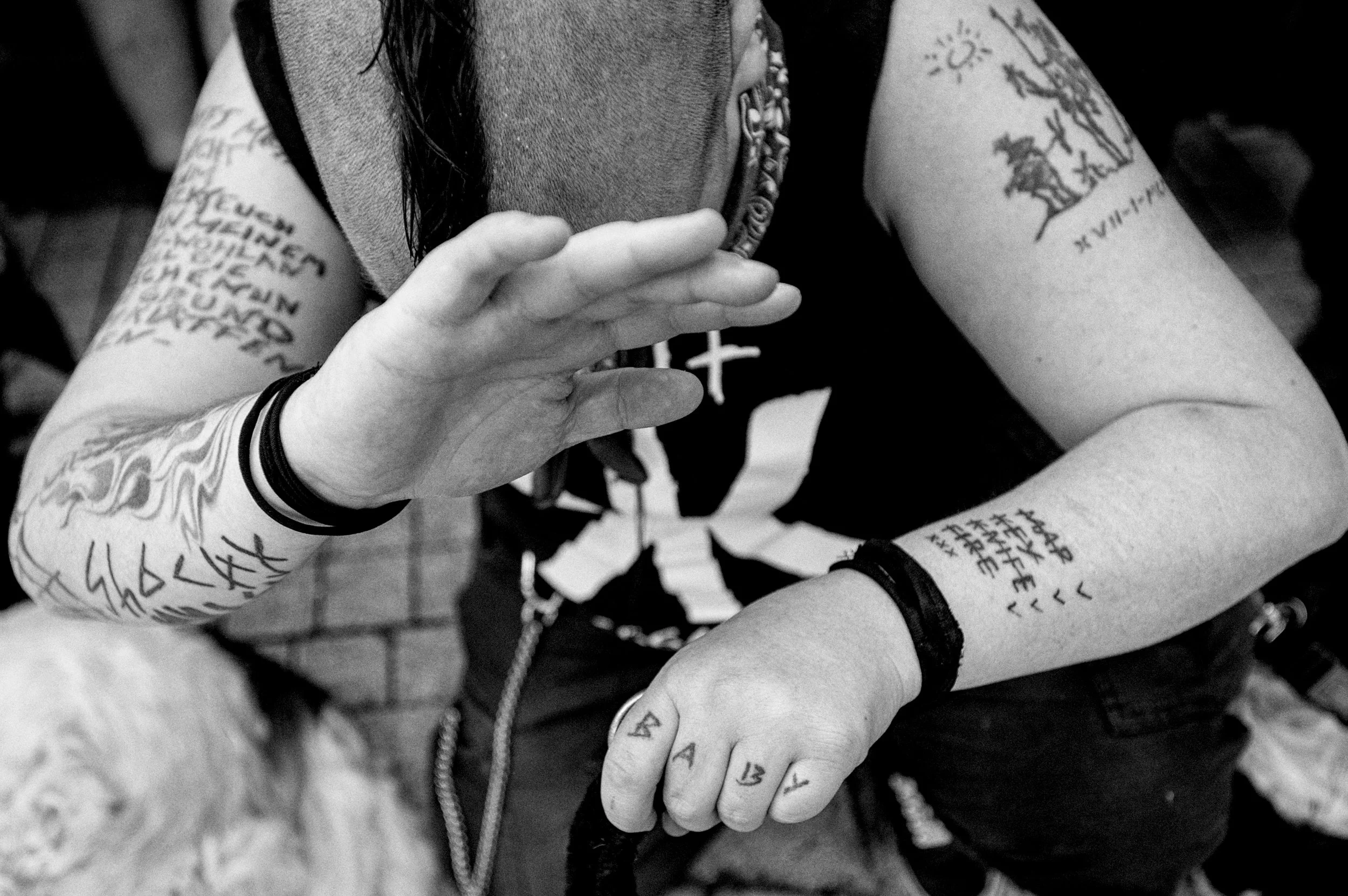 Schwarz-weiße Nahaufnahme einer Person mit vielen Tattoos auf Armen und Händen, die eine Handhaltung zeigt. Die Tattoos enthalten Texte und Symbole, die teilweise sichtbar sind.