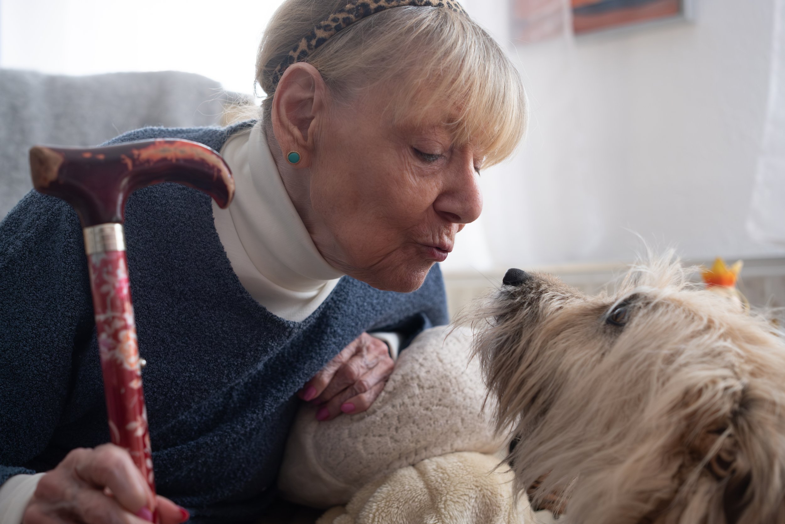 Ältere Frau mit Blümchenschirm lehnt sich zu einem Hund, beide schauen sich liebevoll an.