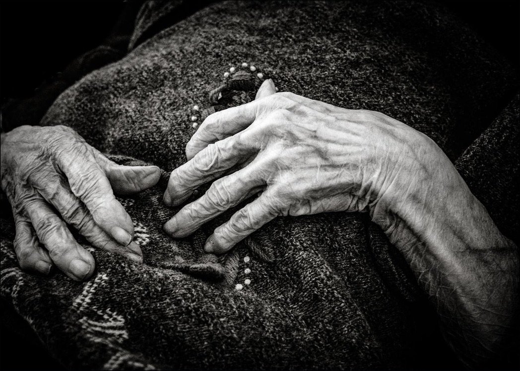 Nahaufnahme von zwei alten Händen, die auf einem dunklen Stoff ruhen, eine mit einer Perlenkette