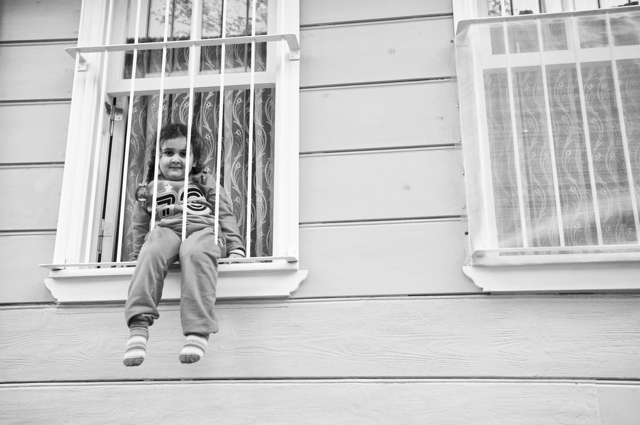 Ein junges Mädchen sitzt an ausgestelltem Fenster in einem Haus, umgeben von Fensterläden und Gardinen, in schwarz-weiß.