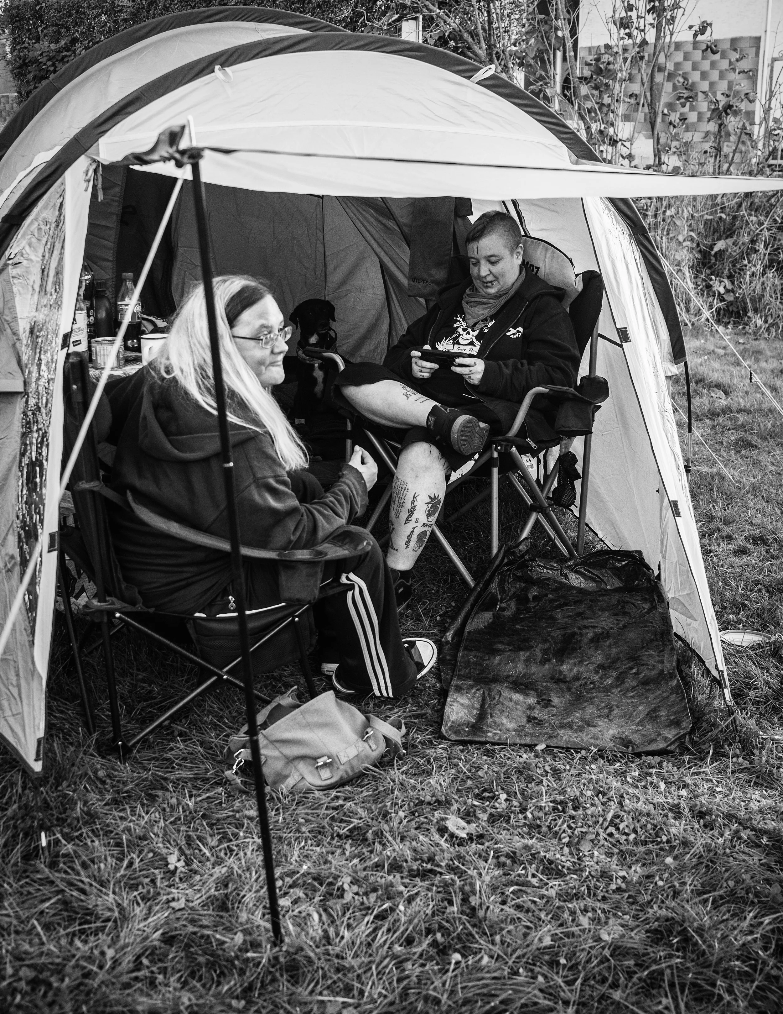 Zwei Menschen sitzen in einem Zelt im Freien, eine auf einem Campingstuhl, beide unterhalten sich, im Hintergrund ein Hund und ein Garten.