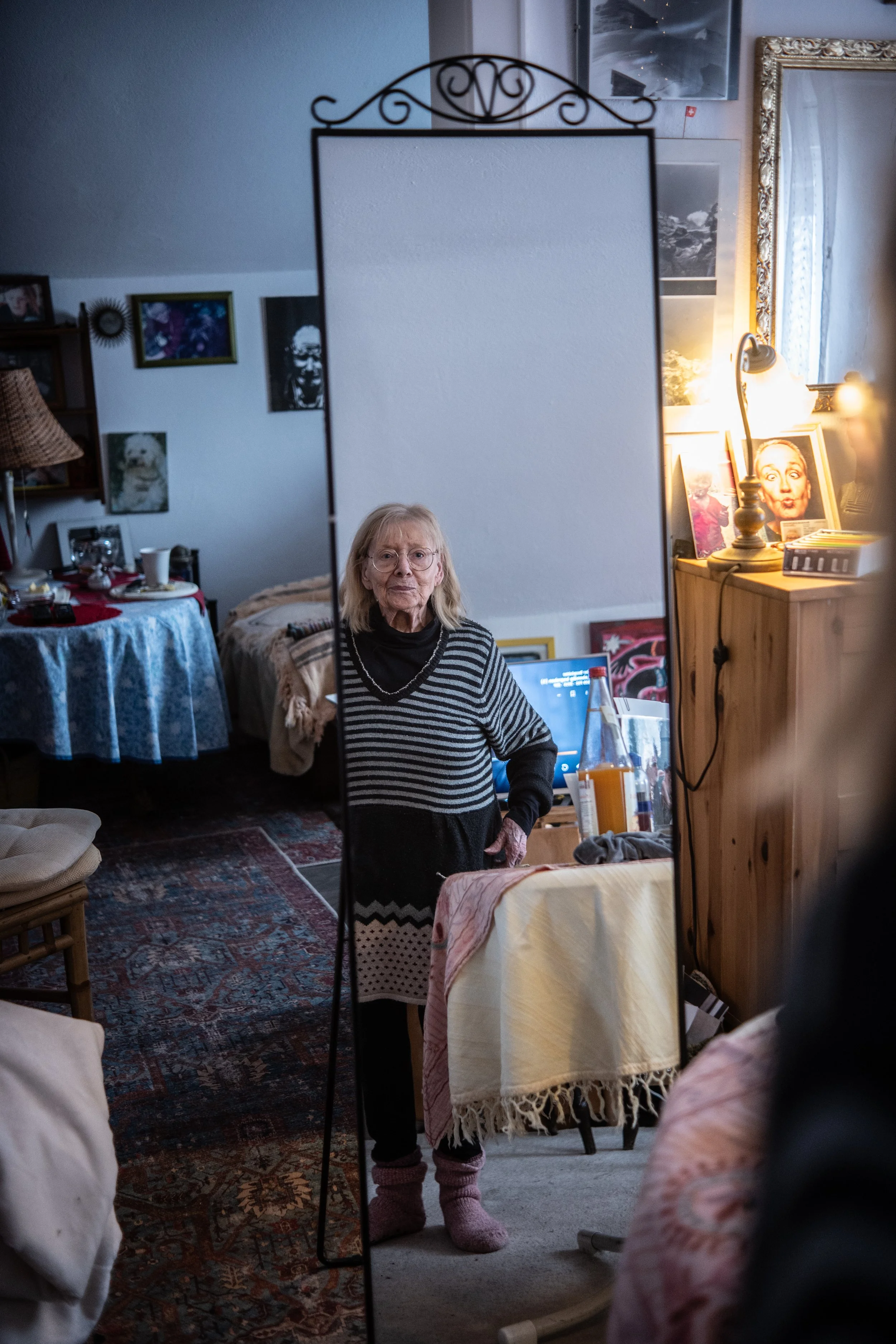 Ältere Frau mit blonden Haaren und Brille, die in einem Wohnzimmer vor einem Spiegel steht.