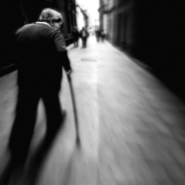 Ein älterer Mensch mit Gehstock geht auf einer Stadtstraße, im Hintergrund weitere Personen.