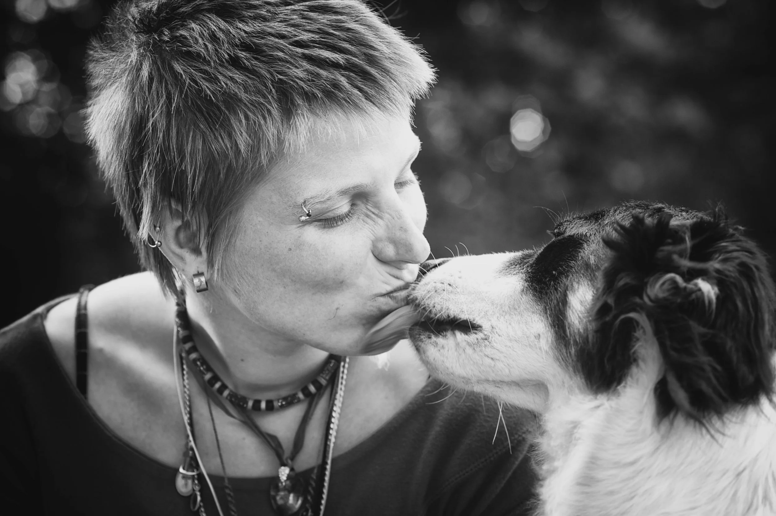 Eine Person mit kurzhaarfrisur und mehreren Piercings küsst einen Hund auf die Nase, im schwarzen und weißen Bild.