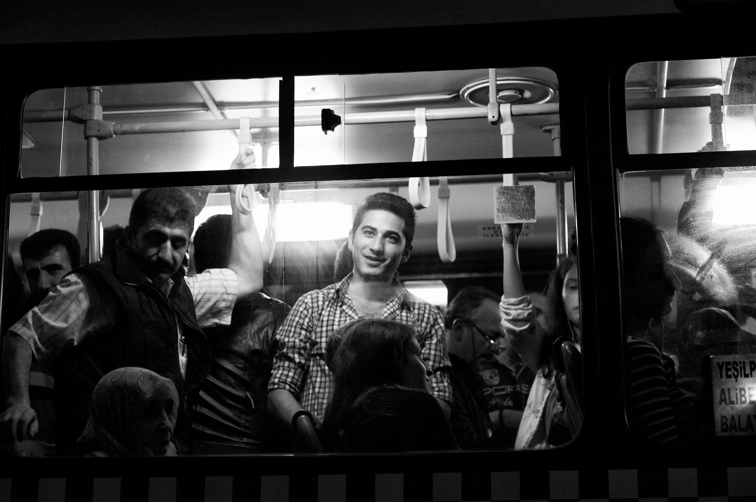 Menschen in einem Bus, einige sitzen, andere stehen, im Schwarz-Weiß-Bild, mit Griffen und Haltestangen.