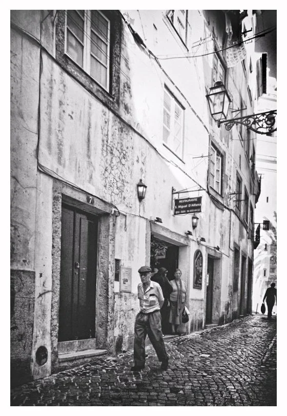 Schmale Kopfsteingasse in einer alten Stadt mit vier Personen, darunter ein älterer Mann mit Brille, und einem Restaurant-Schild