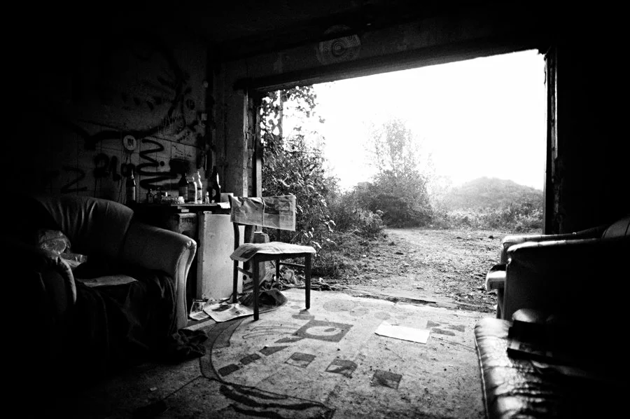 Innenraum eines verlassenen, dunklen Gebäudes mit Sofas, einem Tisch, einem Stuhl und einem offenen Durchgang zur Natur draußen.