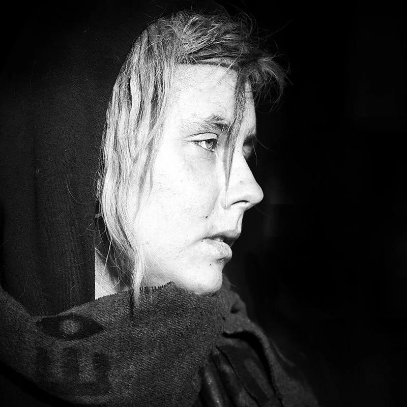 Schwarz-weiß Porträt einer jungen Frau in Seitenansicht mit nassem, blondem Haar und dunklem Hoodie, gegen einen schwarzen Hintergrund.