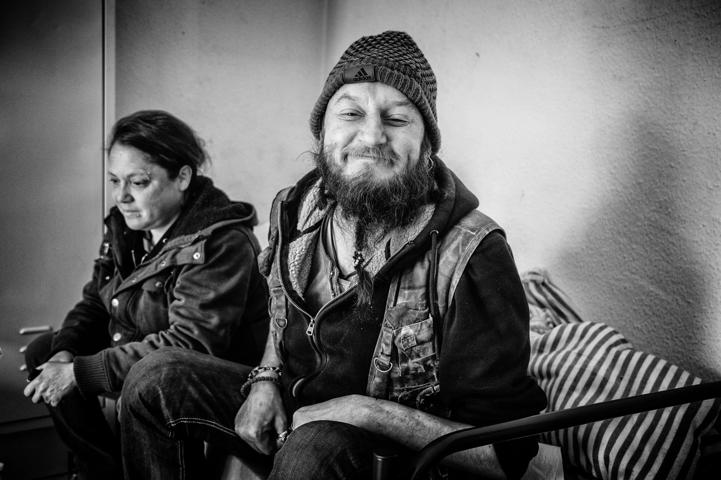 Ein lächelnder Mann mit Strickmütze und Vollbart sitzt in einer Einrichtung, neben ihm eine ernst wirkende Frau. 