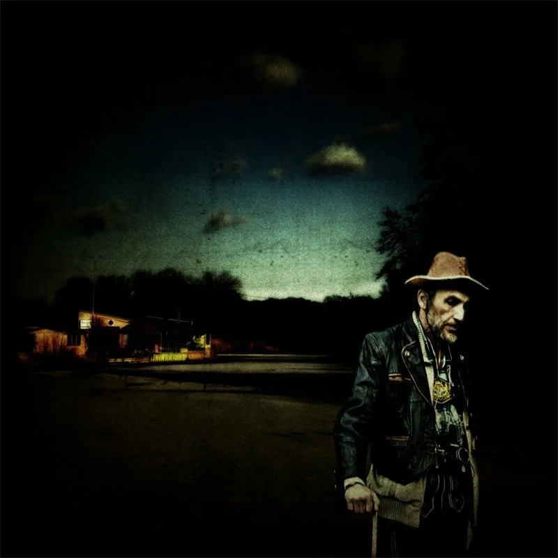 Ein alter Mann mit Hut und Spazierstock steht nachts im Freien, im Hintergrund sind Häuser und dunkle Bäume, der Himmel ist bewölkt.