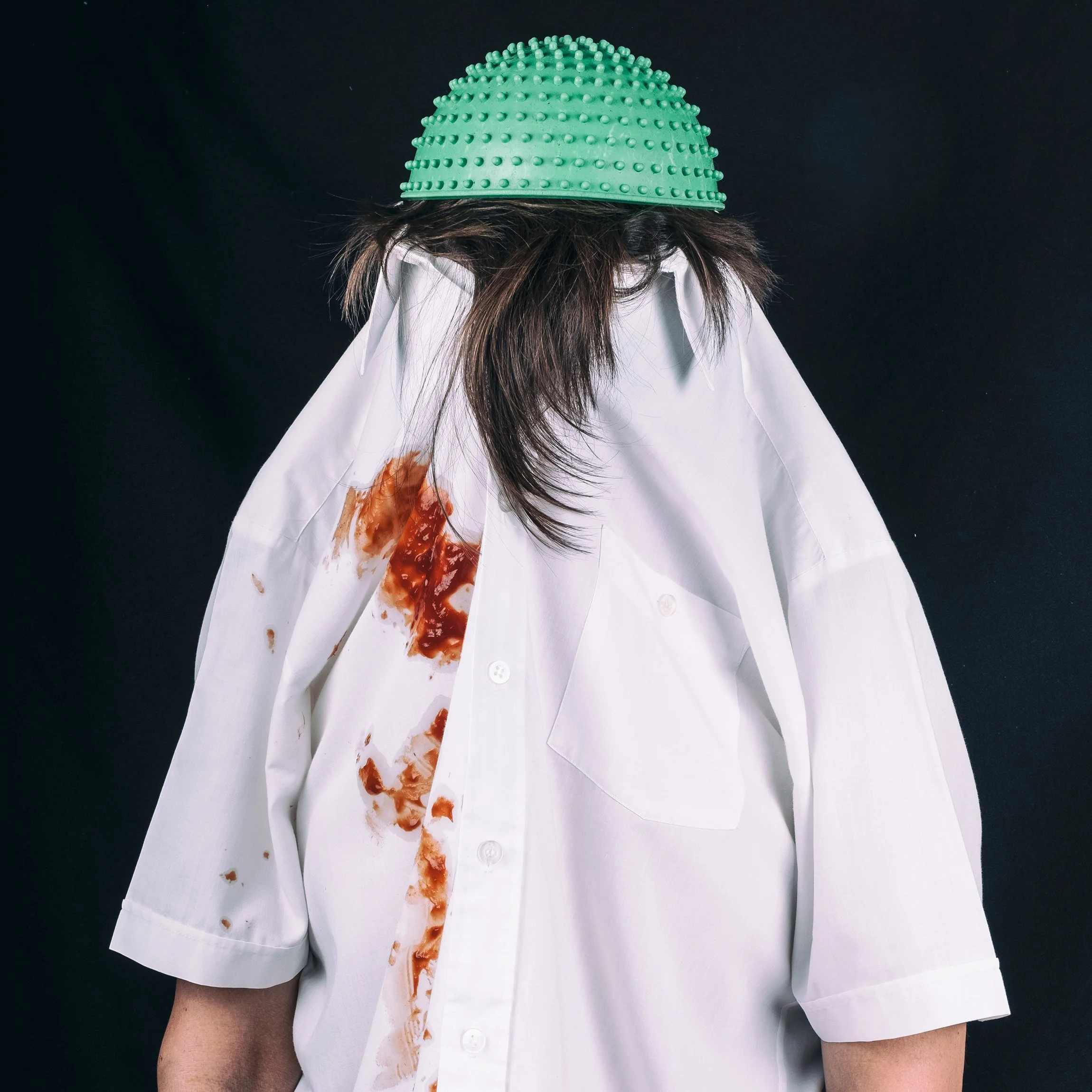 Eine Person trägt eine weiße Hemd, das am linken Oberarm mit Ketchup verschmutzt ist, und einen grünen Schleimhut auf dem Kopf.