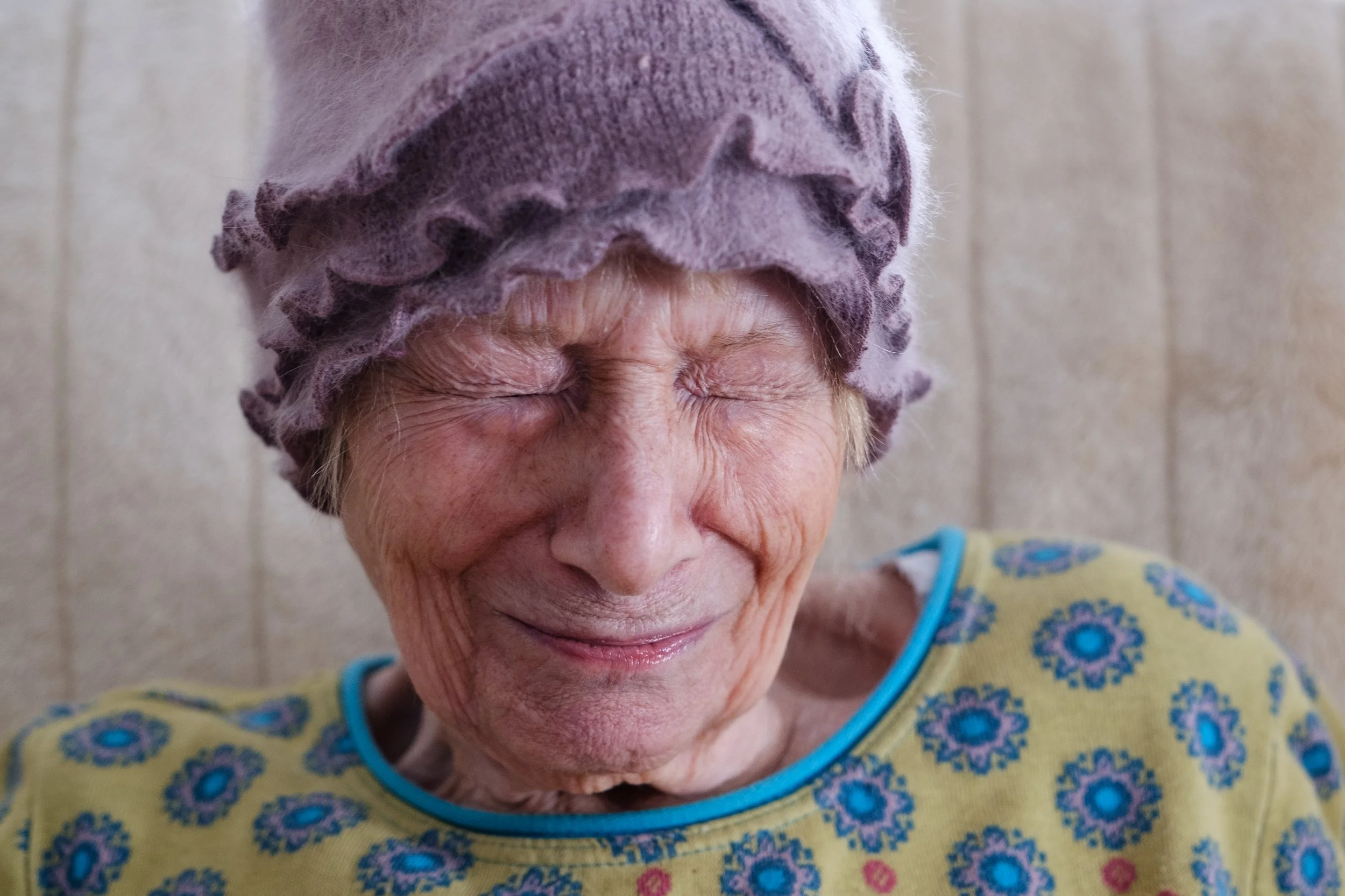 Lächelnde ältere Frau mit violettem Haarschutz und bunter Blümchen-Pullover, frontal fotografiert.