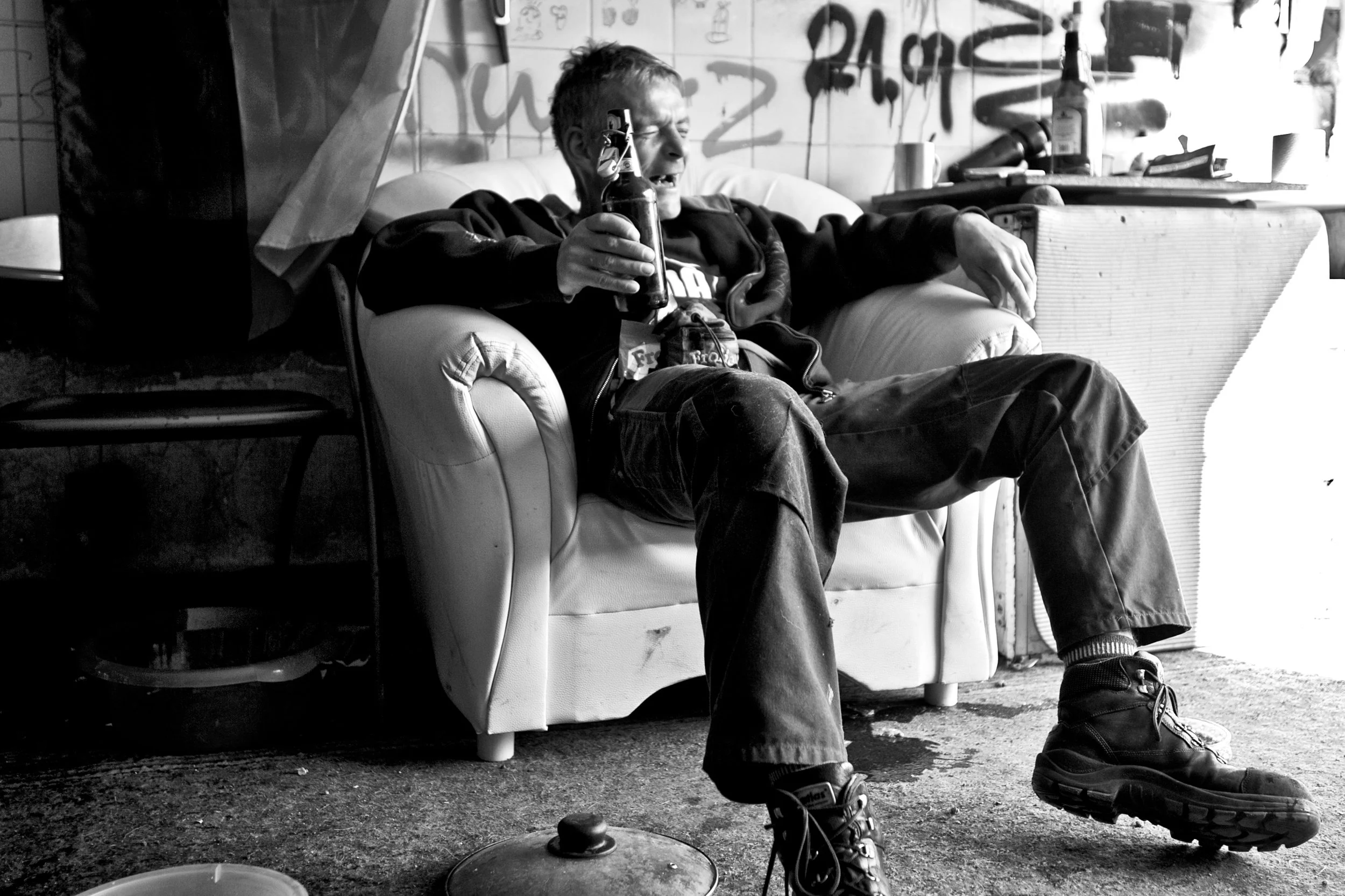 Ein junger Mann sitzt auf einem Sofa in einem ungeordneten Raum, hält eine Bierflasche in der Hand und scheint zu lachen oder zu sprechen. Der Raum hat Graffiti an der Wand im Hintergrund, und es gibt verschiedene Gegenstände auf einem Tisch und im R