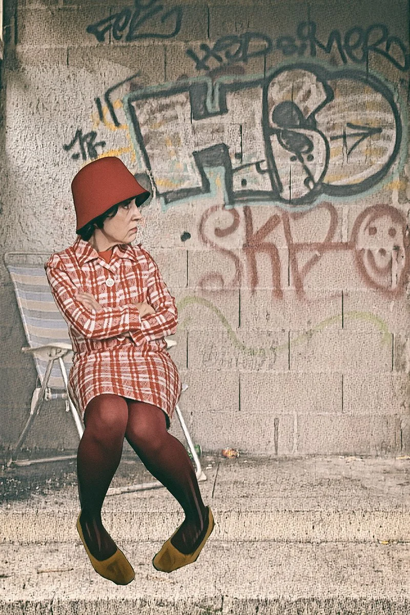 Eine Frau sitzt mit verschränkten Armen vor einer mit Graffiti bedeckten Wand, trägt einen roten Hut, ein rotes kariertes Kleid, rote Strumpfhosen und gelbe Schuhe.