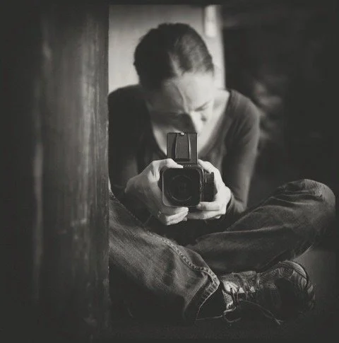 Schwarz-weiß-Foto einer Person, die in einer sitzenden Position ein Kamerabildschirm zeigt, während sie im Spiegel fotografiert wird.