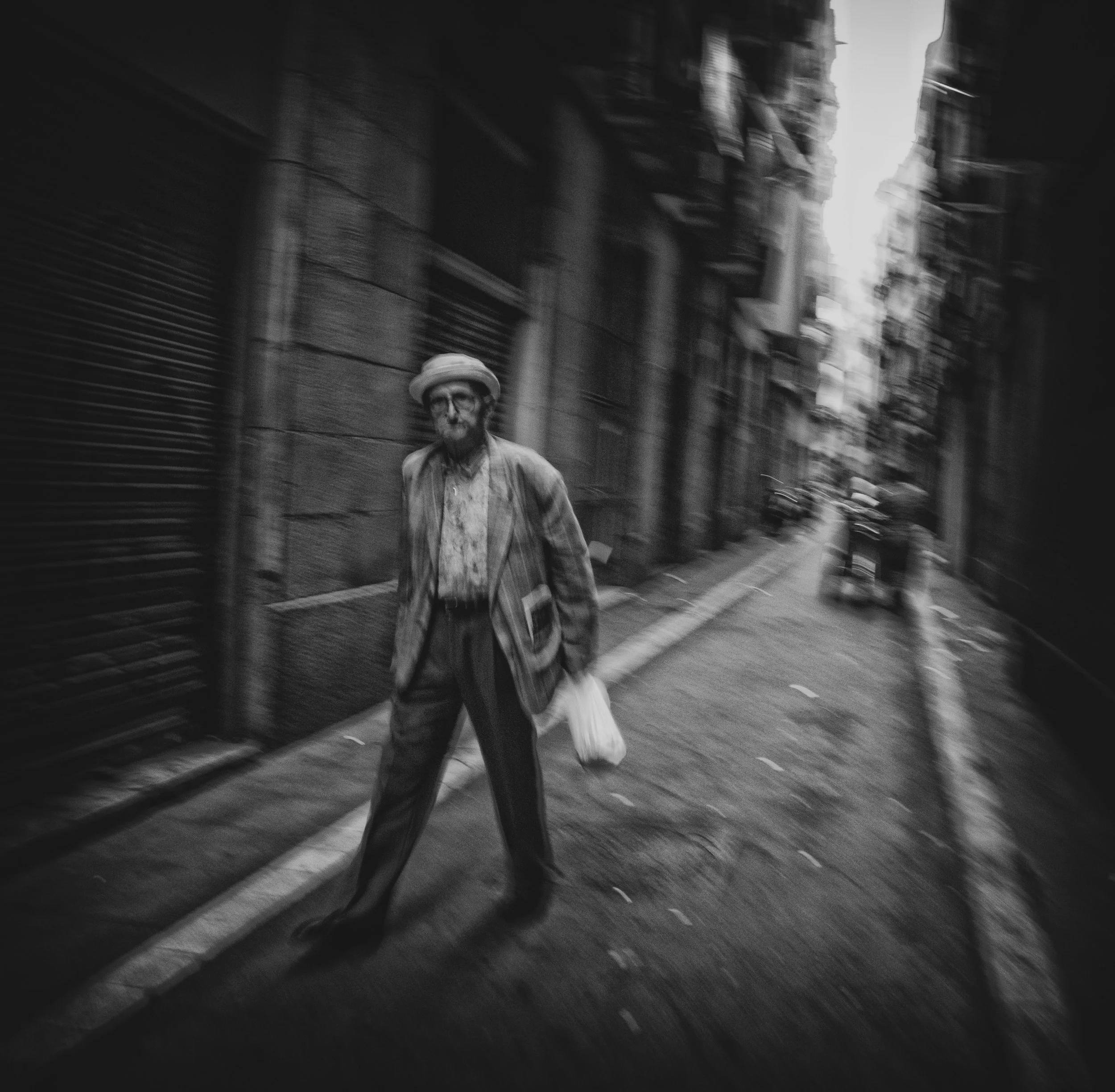 Ein älterer Mann in gestreifter Kleidung und Hut steht im Vordergrund auf einer engen, urbanen Gasse in Schwarz-Weiß. Er hält eine kleine Tüte in der Hand. Die Straße ist schmal mit geschlossen wirkenden Geschäften und einigen Fahrzeugen im Hintergru