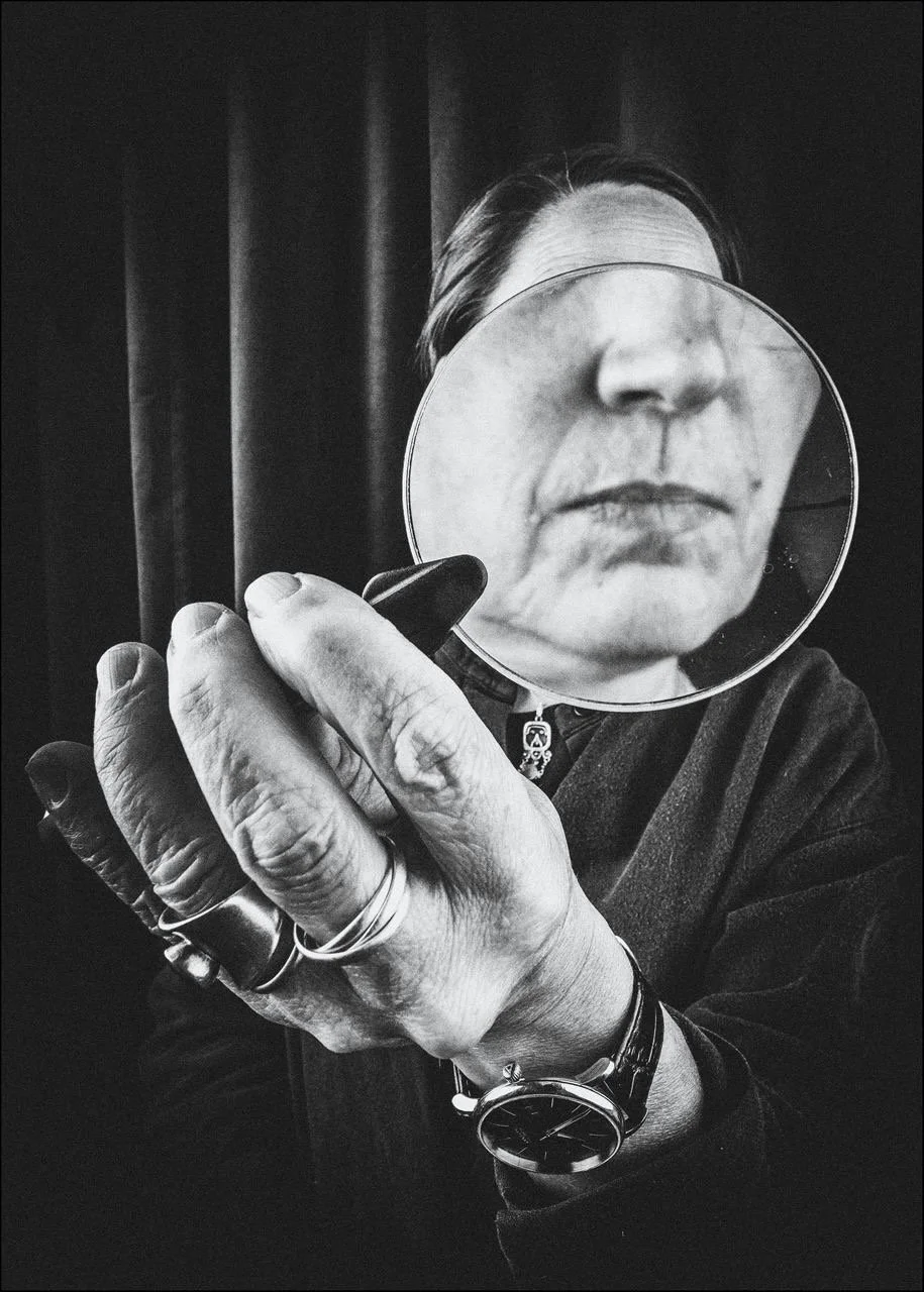 Eine Person hält einen Handspiegel vor das Gesicht, der das Gesicht im Spiegel anzeigt, der eine Nahaufnahme des Mundes und der Nase zeigt; das Bild ist in Schwarzweiß.