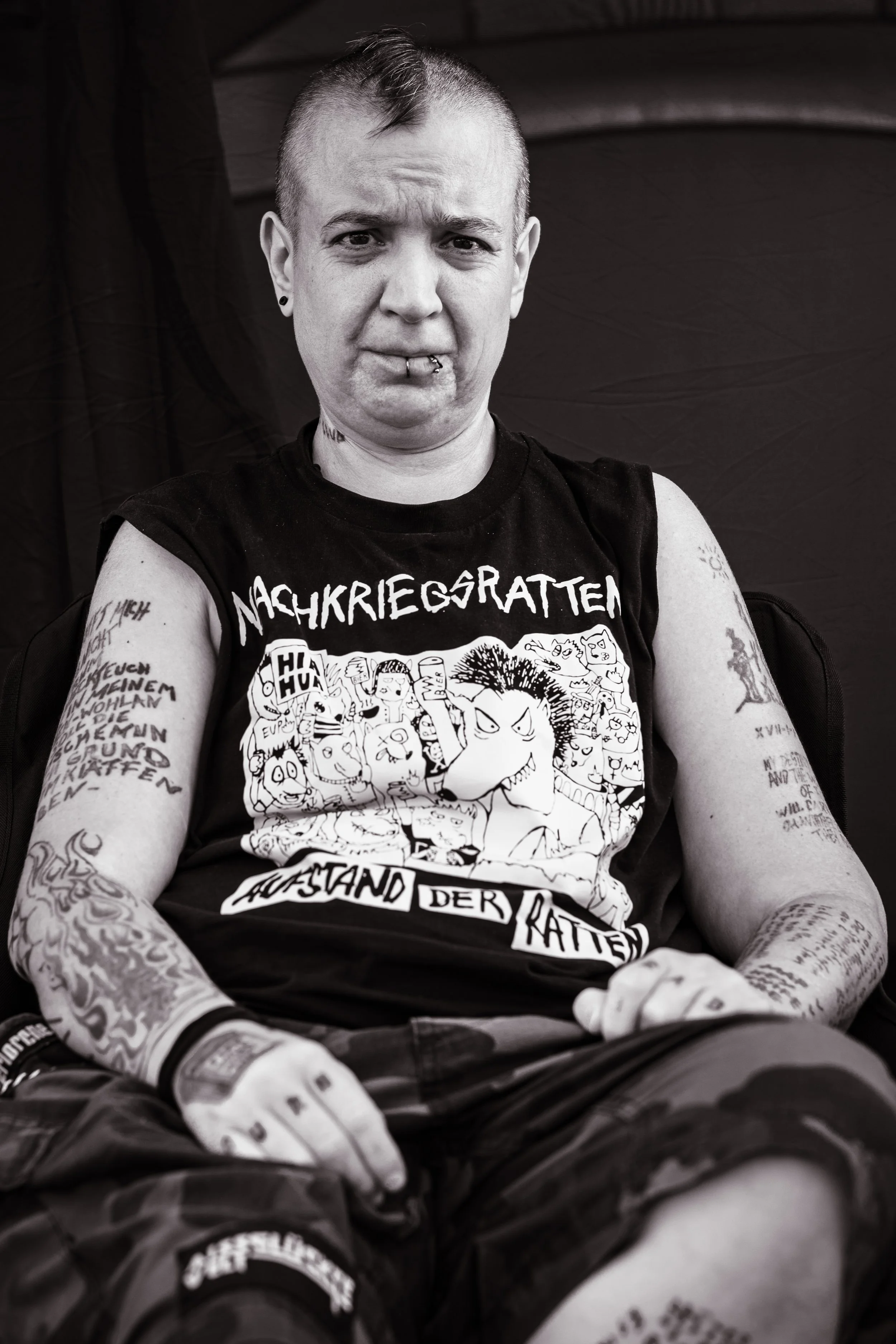 Schwer tätowierte junge Person mit Piercings trägt ein schwarzes T-Shirt mit Cartoon-Designs und deutschen Texten, sitzt entspannt.