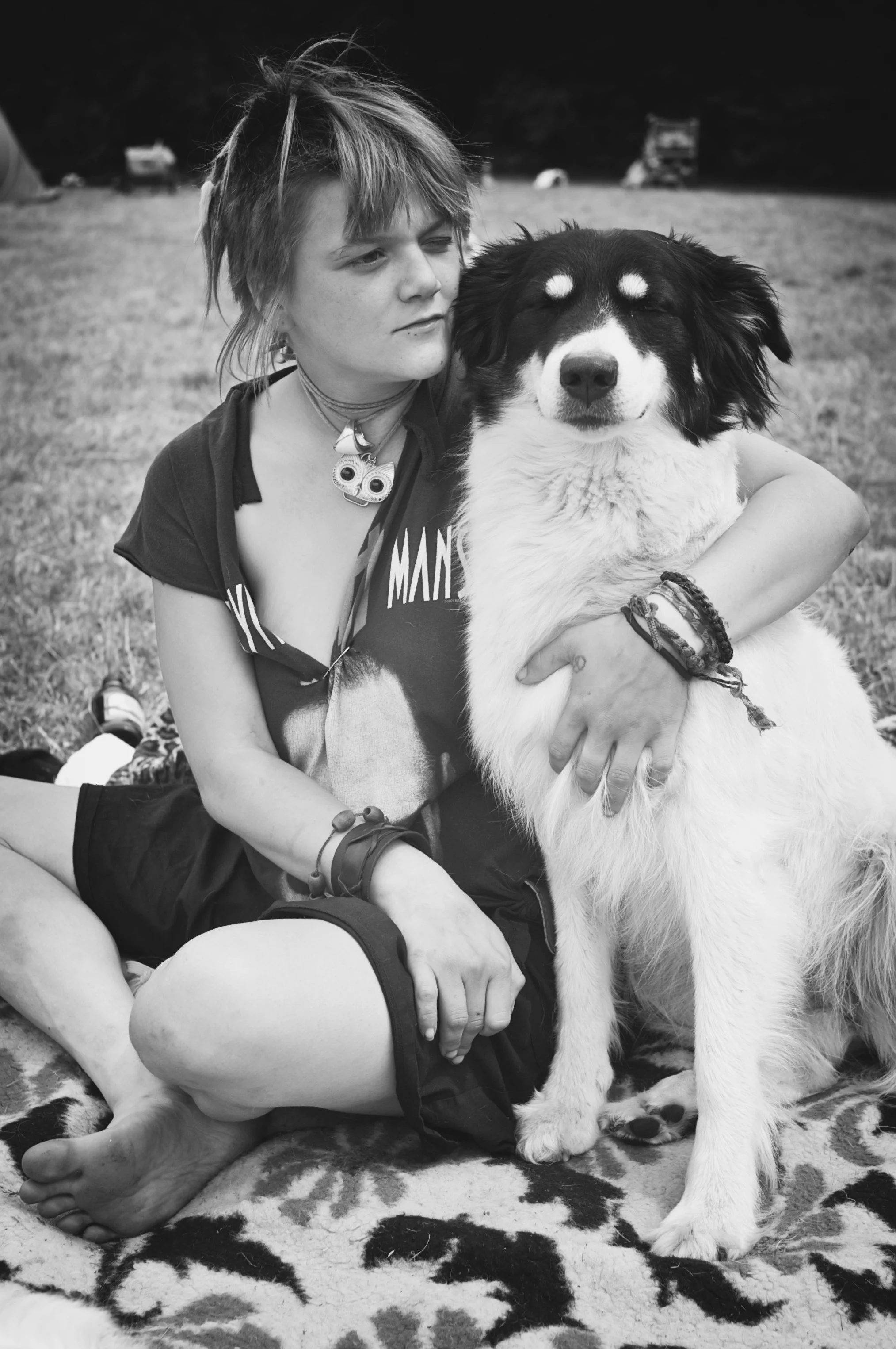 Ein Mädchen sitzt auf dem Boden, das Bein angezogen, während sie einen Hund umarmt, beide in einer Feldlandschaft. Das Mädchen trägt Armbänder und eine Halskette, hat kurze Haare und sieht nachdenklich. Der Hund sitzt entspannt mit geschlossenen Auge