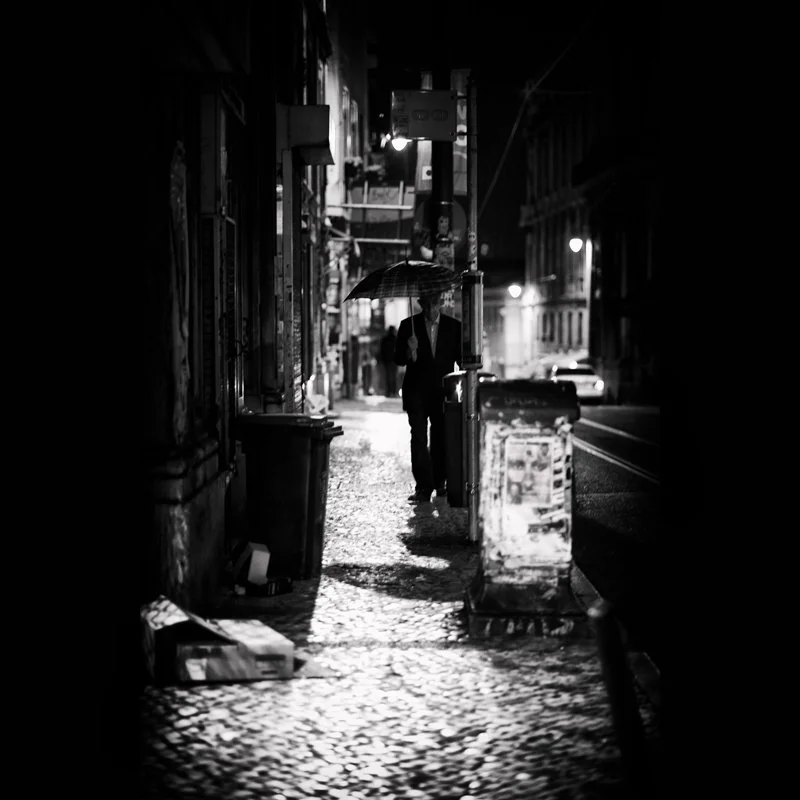 Ein Mann mit Regenschirm läuft nachts auf einer nassen Straßeninsel, im Hintergrund leuchten Straßenlaternen und Fahrzeuge, Schwarz-Weiß-Aufnahme.