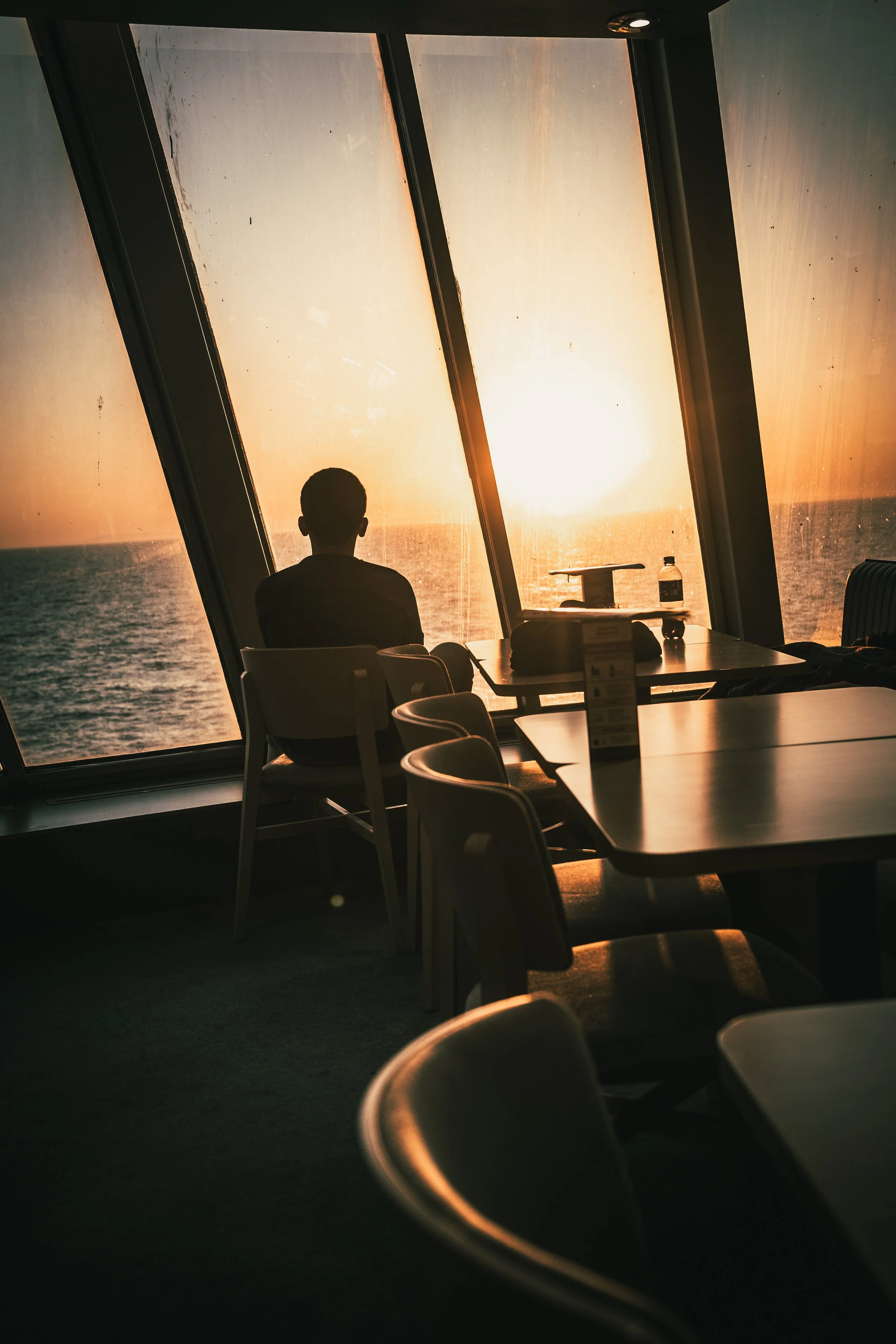 Ein Mensch sitzt in einem Raum mit großen Fenstern, die den Sonnenuntergang über dem Meer zeigen, während die Sonne im Hintergrund untergeht.