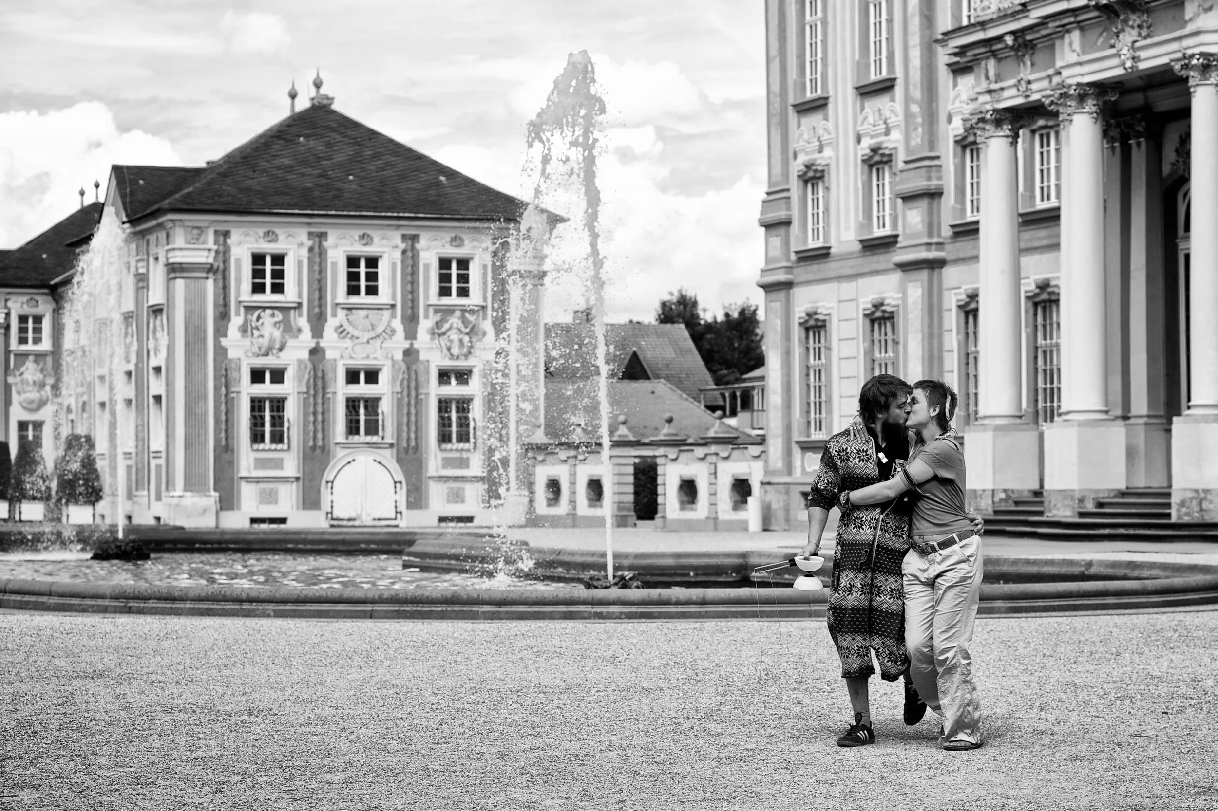 Zwei Menschen küssen sich vor einem historischen Gebäude mit Springbrunnen, im schwarzen und weißen Foto