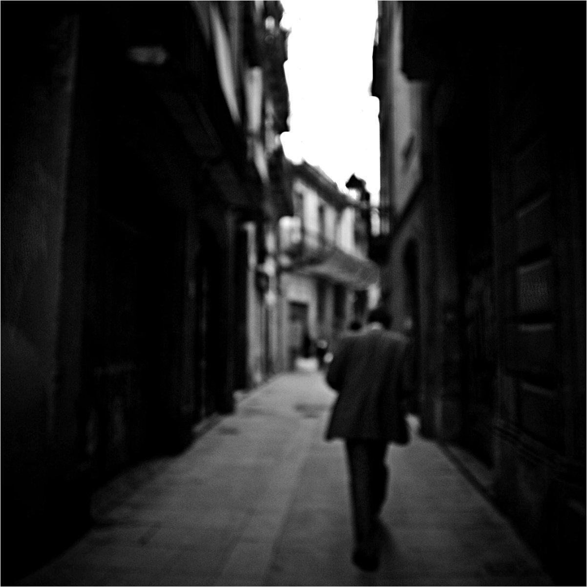 Eine schwarz-weiße Aufnahme eines Mannes, der die Straße entlang geht. Er trägt einen Mantel und hat die Rückseite zur Kamera. Die Gebäude auf beiden Seiten sind alt und schmal, die Szene wirkt urban und ruhig.