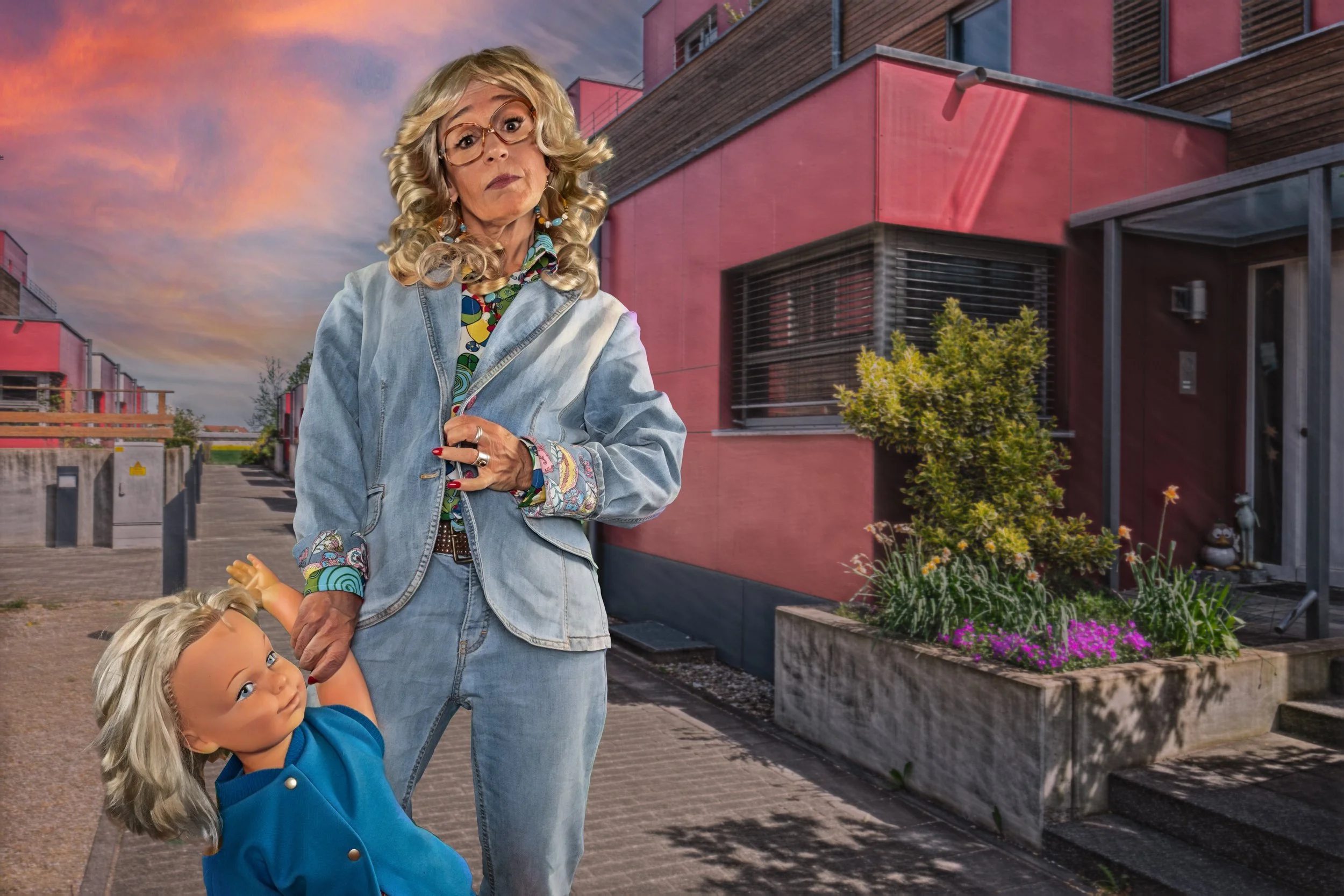 Eine ältere Frau mit langen, lockigen, grauen Haaren und Brille, die eine Jeansjacke trägt, hält die Hand eines kleinen Puppenjahres, das wie ein Kind aussieht. Im Hintergrund sind moderne, pinkfarbige Häuser mit Gartenzubehör und Pflanzen sichtbar, 