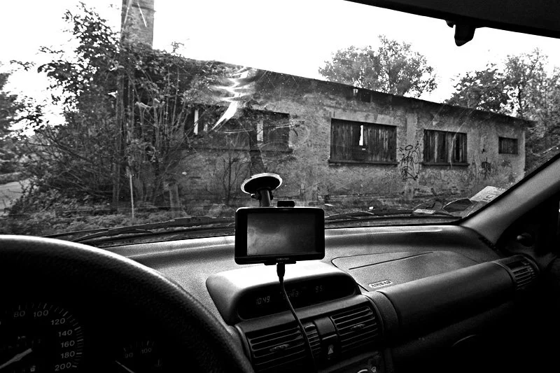 Blick aus dem Inneren eines Autos auf eine verlassene, zerfallene Häuserwand mit Graffiti, umgeben von Bäumen und Pflanzen im Schwarz-Weiß-Foto.