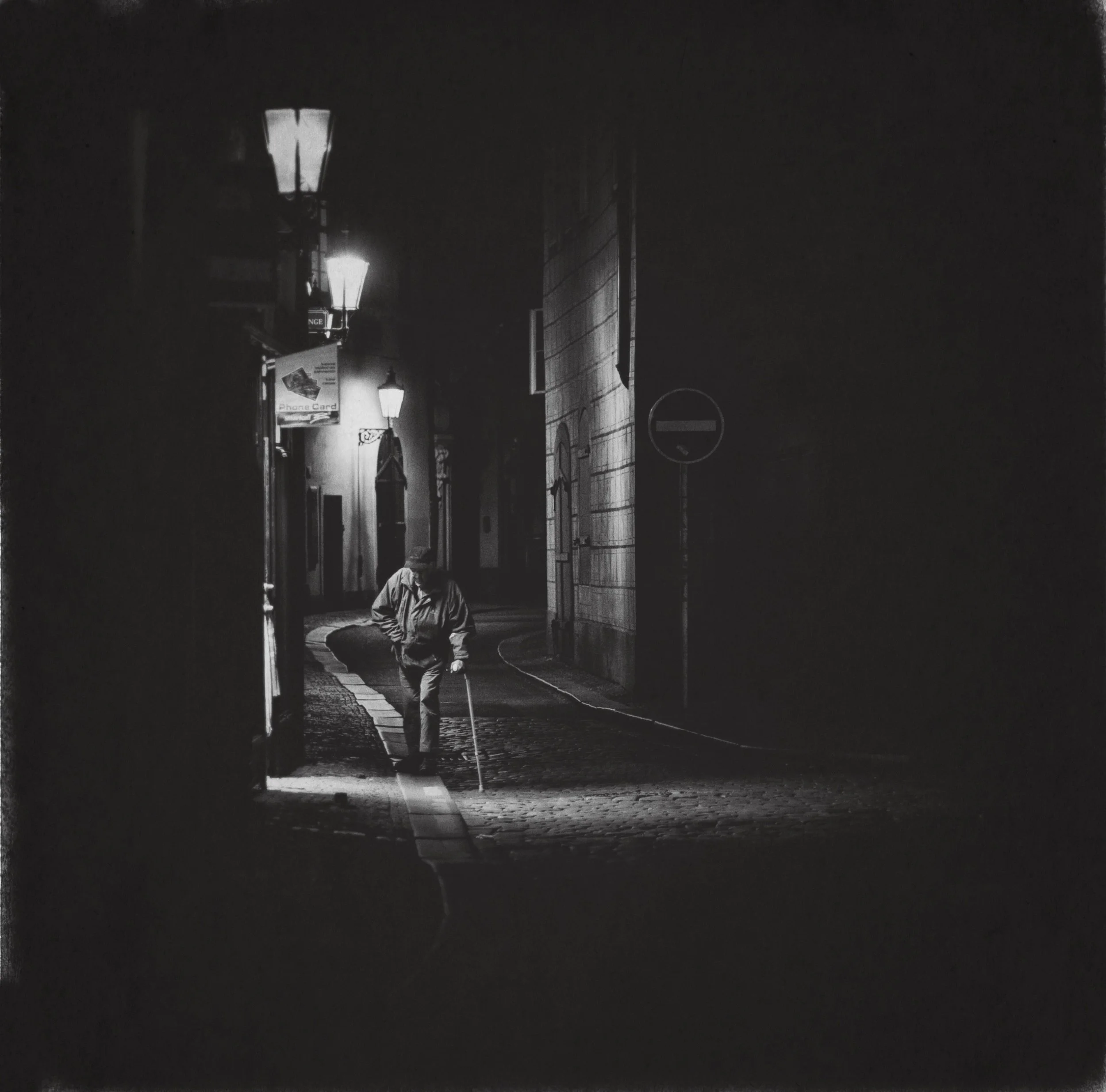 Ein Mensch mit einem Gehstock geht nachts durch eine dunkle, enge Straße, beleuchtet von Straßenlaternen.