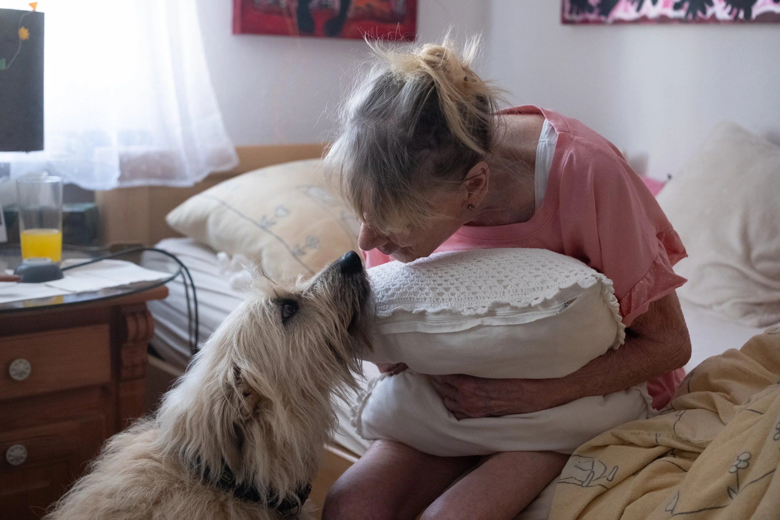 Eine ältere Frau hält ein Kissen und beugt sich mit ihrem Kopf nahe an einen Hund, der auf einem Bett sitzt, in einem Schlafzimmer mit hellen Wänden und Wanddekorationen.