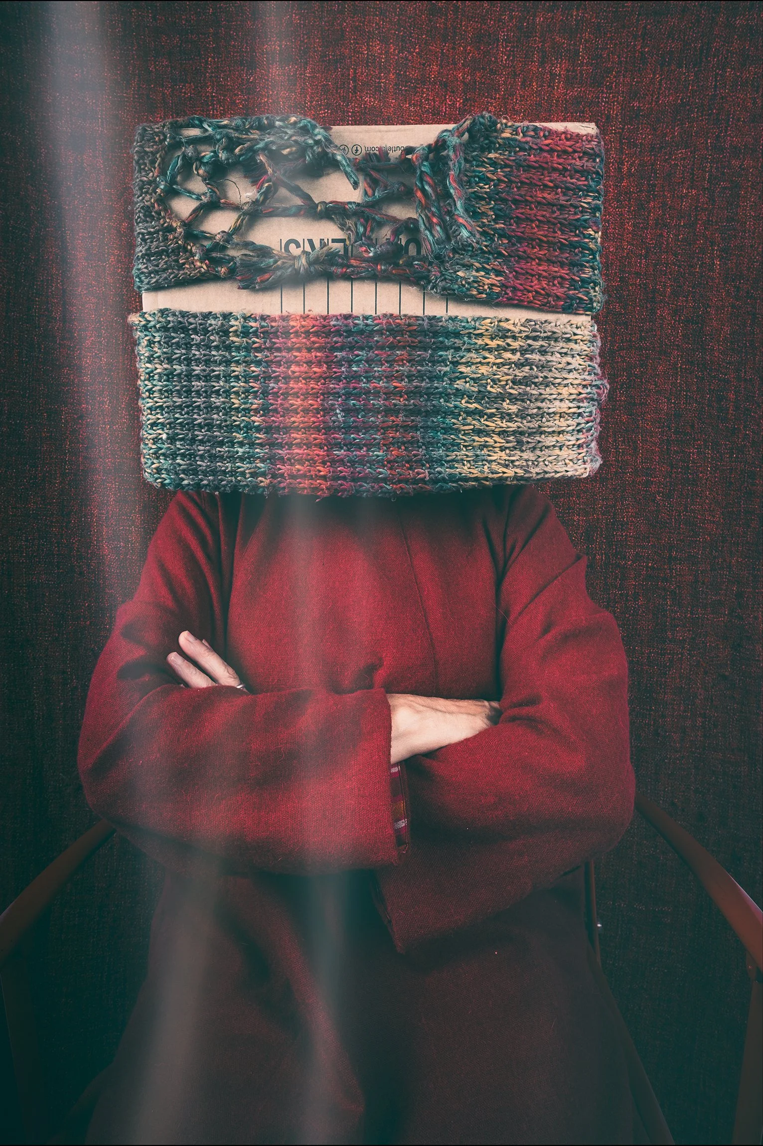 Eine Person sitzt mit verschränkten Armen gegen eine dunkelrote Wand, deren Kopf von einem bunter, gestrickten Schal bedeckt ist. Die Person trägt einen dunkelroten Mantel.