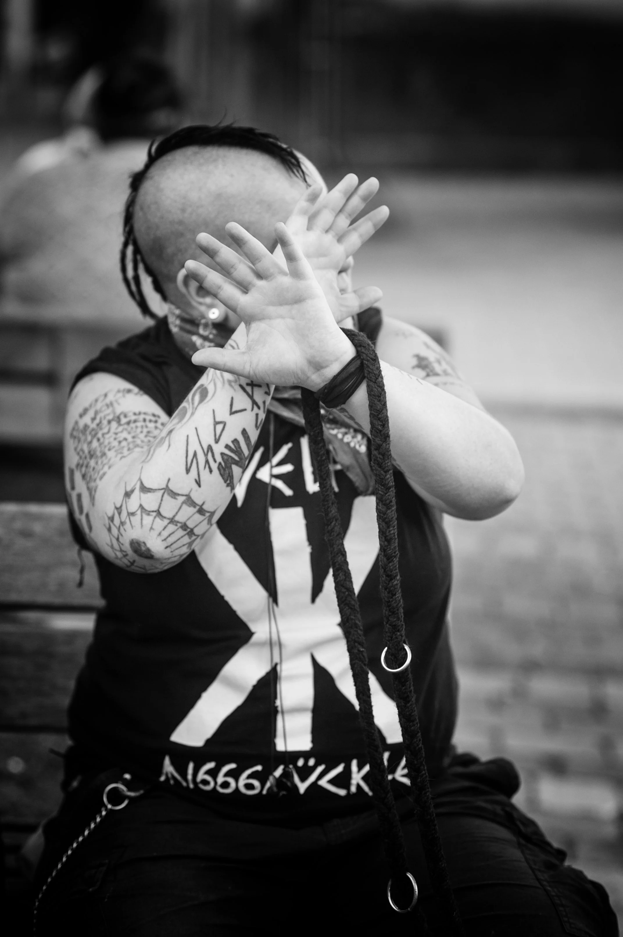 Schwarz-weiß-Foto einer Person mit Tattoos, die die Hände vor das Gesicht hält. Die Person trägt ein T-Shirt mit einem Union Jack-Design. Der Hintergrund ist unscharf und zeigt eine Holzbank.