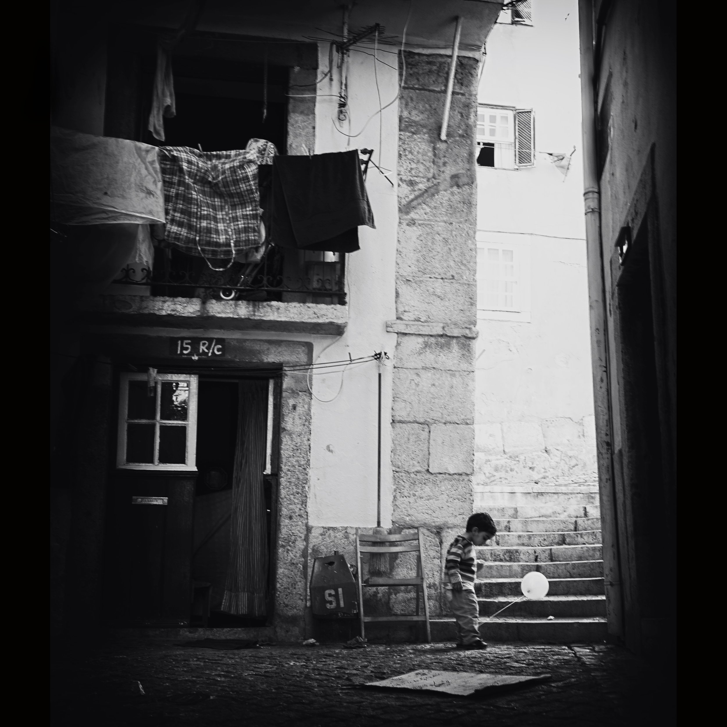 Ein Junge, der mit einem Ballon in einer engen, dunklen Gasse spielt, mit Treppen im Hintergrund und Wäsche, die an einer Leine hängt, typisch für eine alte europäische Stadt.