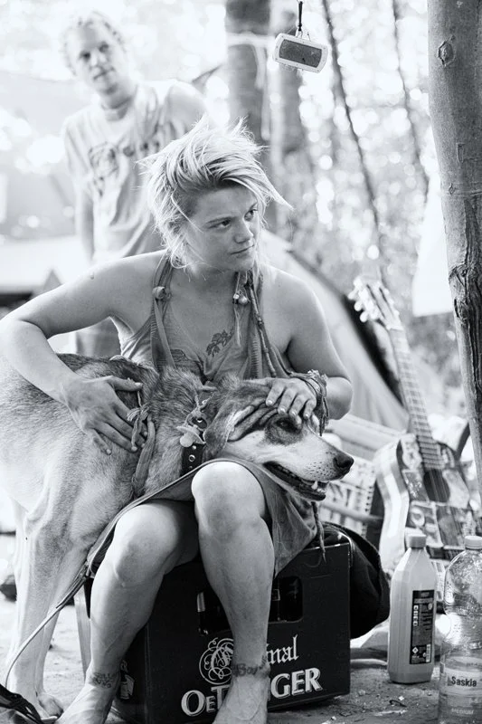 Junge Frau mit kurzer, unordentlicher Frisur streichelt einen großen Hund, während ein Mann im Hintergrund steht. Im Freien, mit mehreren Gegenständen und einer Gitarre im Hintergrund.