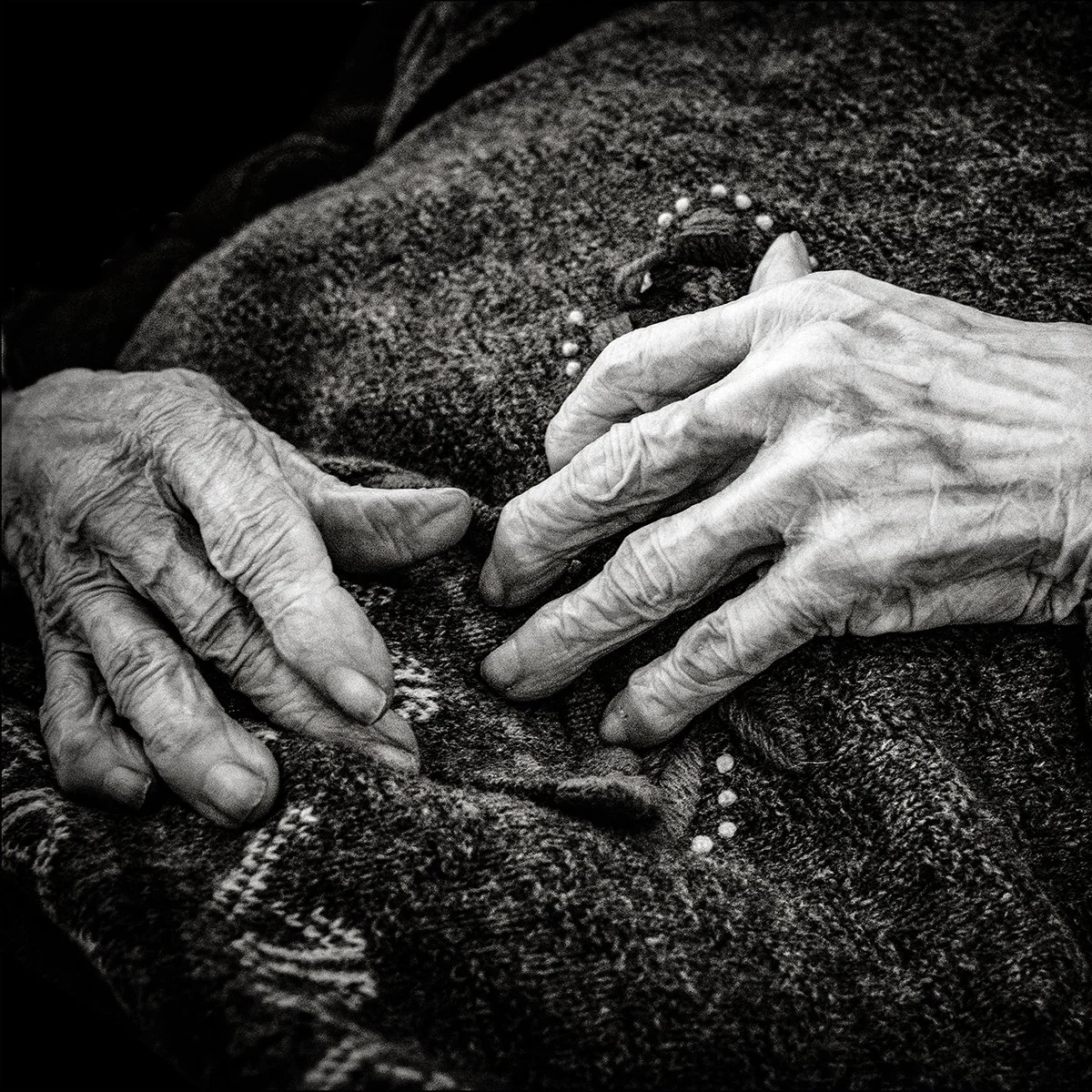 Ein schwarz-weiss Bild von zwei knochigen und altgewordenen Händen auf einer teilweise bestickten Decke