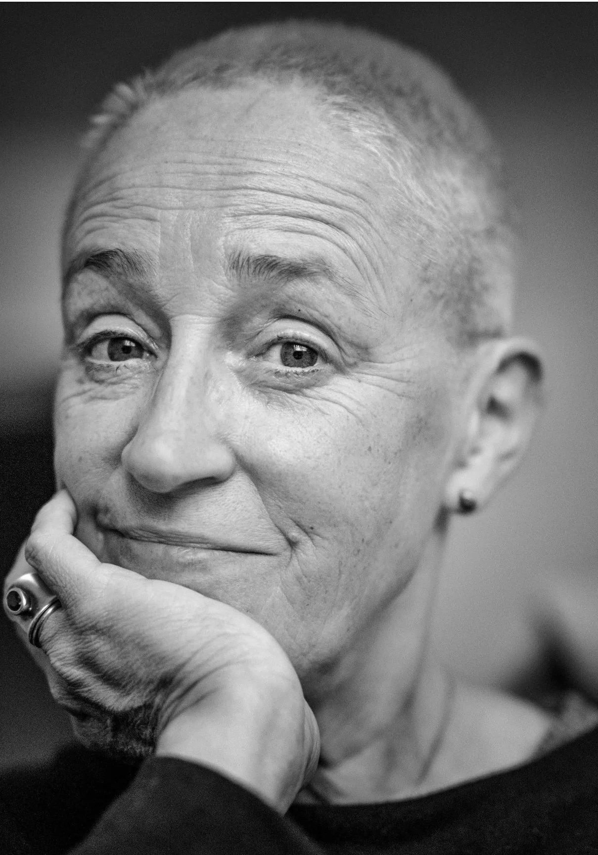 Schwarz-Weiß-Foto einer lächelnden älteren Frau, die die Hand an ihr Kinn legt, mit kurz grauhaarigem Haar, einem kleinen Ohrring und einem schwarzen Oberteil.