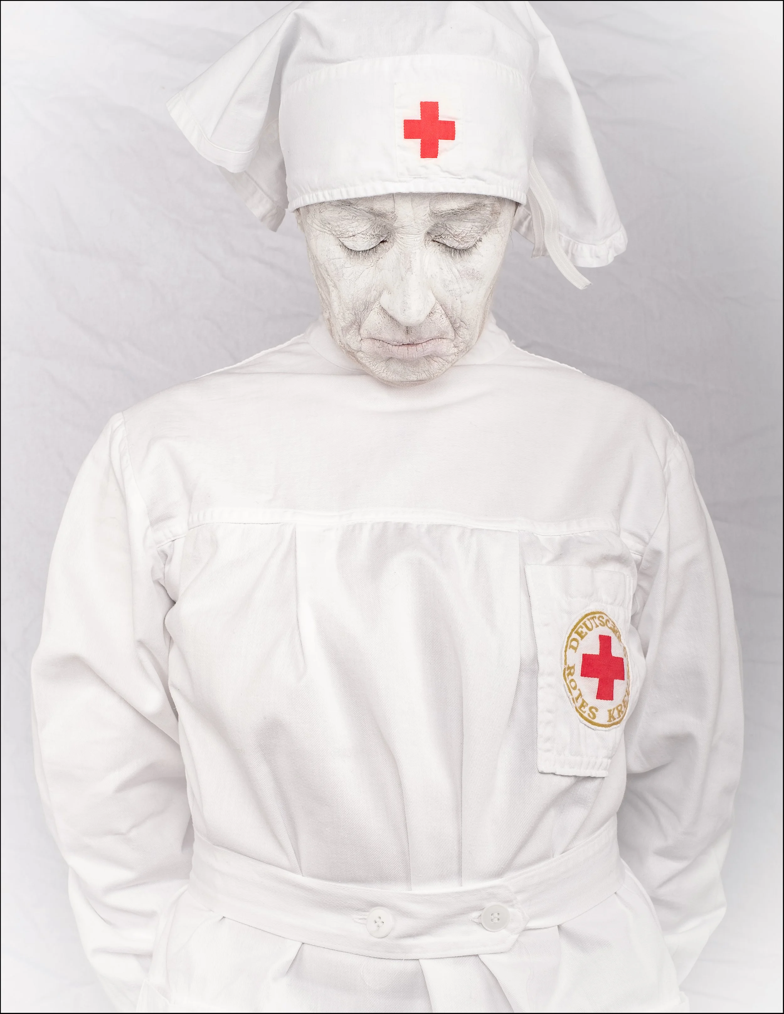 Person in Krankenschwester-Uniform mit weißem Gesicht und geschlossenen Augen, trägt eine weisse Schwesternhaube mit rotem Kreuz und steht vor weißem Hintergrund. Der Kopf ist gesenkt, die Hände sind hinter dem Rücken verschränkt.