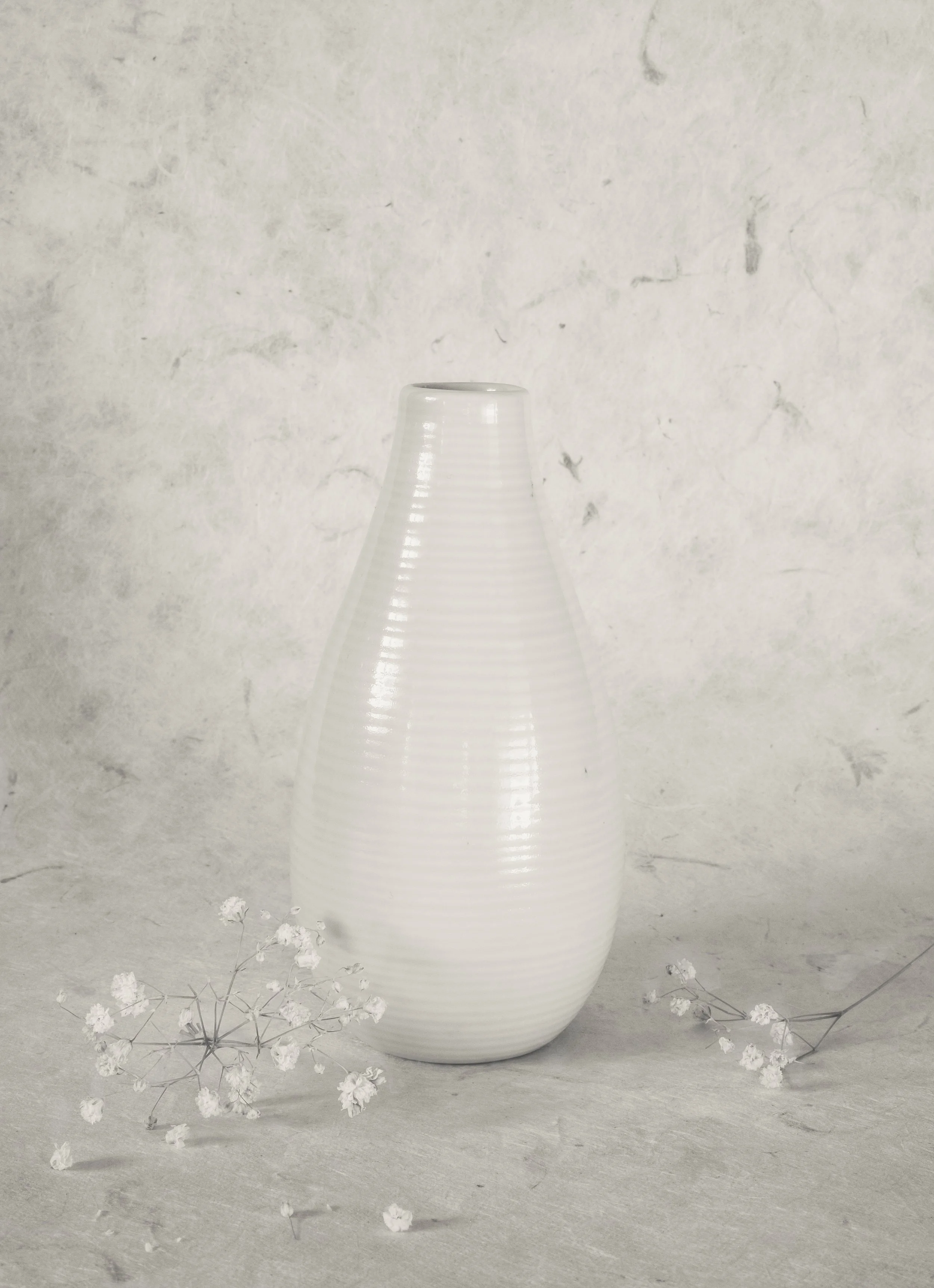 Eine weiße Keramikvase mit geripptem Design und einer ovale Öffnung auf der Oberseite, auf einem hellgrauen Untergrund, flankiert von kleinen weißen Blumen.