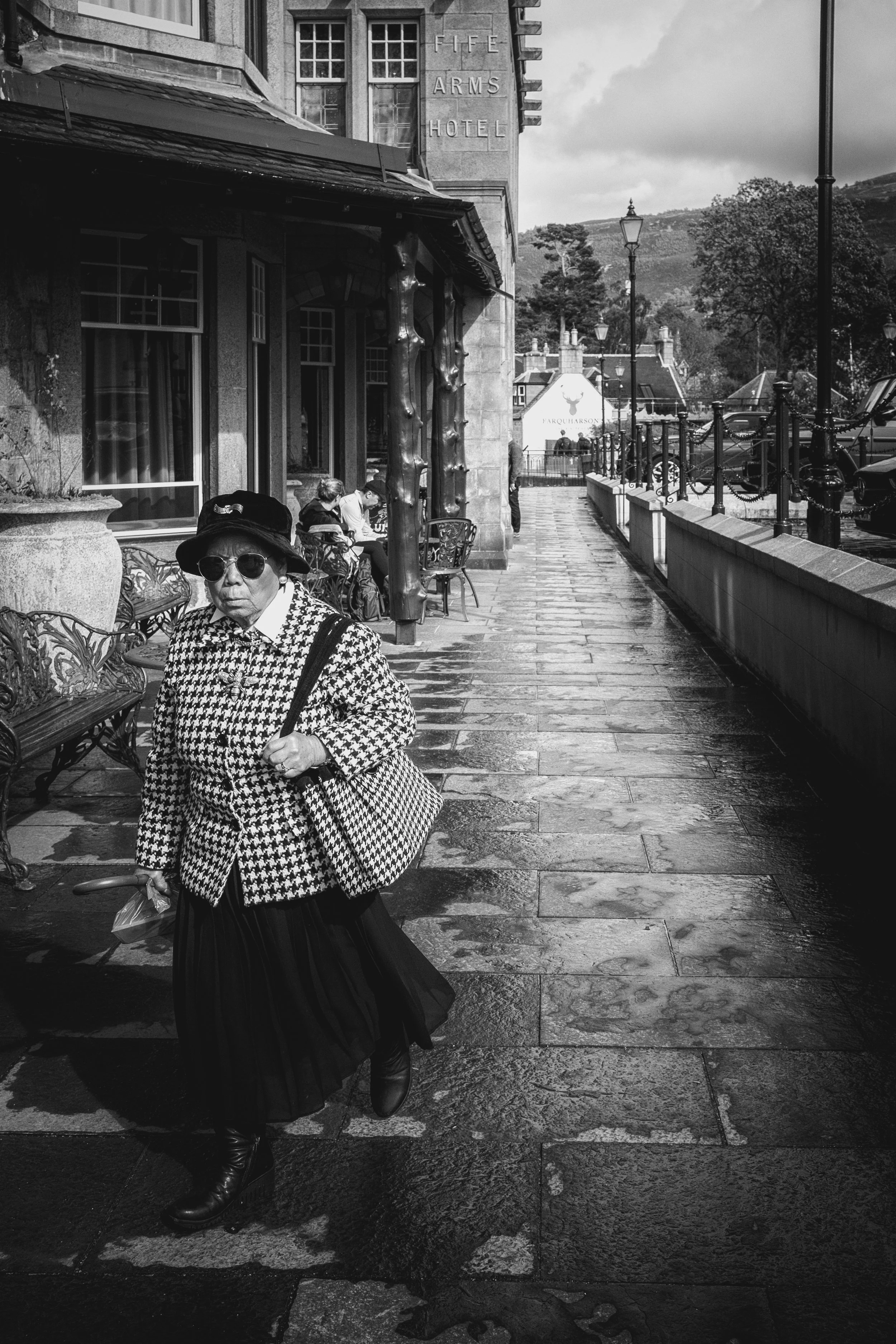 Eine ältere Frau trägt ein schwarz-weißkariertes Mantel und eine dunkle Mütze, läuft auf einer nassen Kopfsteinpflasterstraße vor einem Hotel namens 'Fife Arms' in einer Stadt mit einigen Menschen im Hintergrund.
