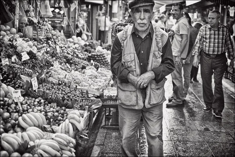 Mann geht auf einem Markt an einem Obst- und Gemüsetresen vorbei, der mit verschiedenen Früchten wie Melonen, Trauben und Beeren gefüllt ist, im Hintergrund andere Marktbesucher.
