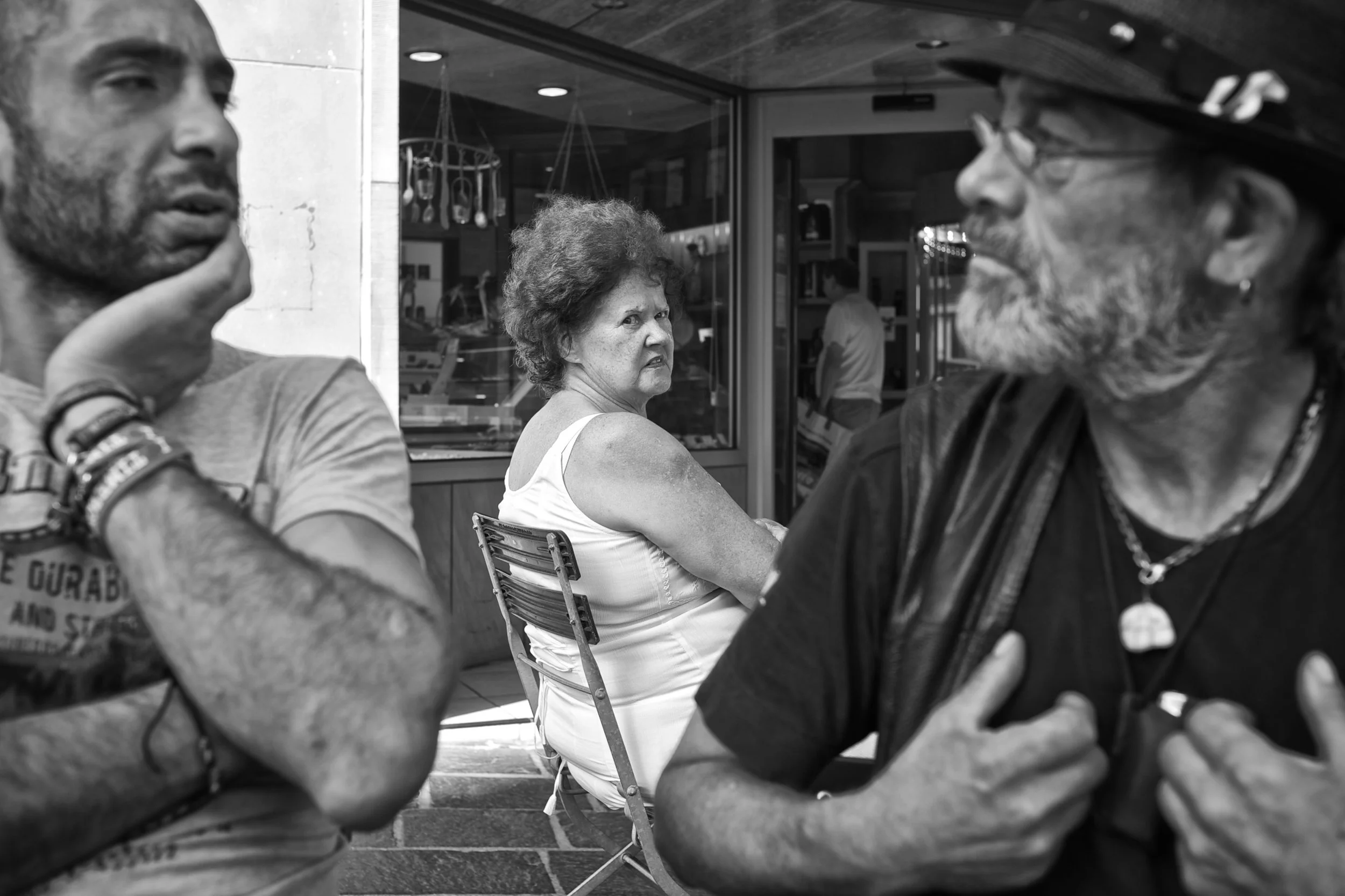 Drei Menschen im Gespräch vor einem Café, eine Frau schaut empört, während zwei Männer sich abwechselnd anschauen.
