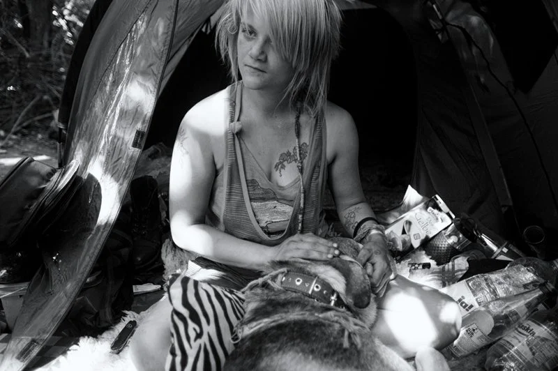 Junge Frau mit langen, gestylt Haaren, sitzend vor einem Zelt, umgeben von Campingausrüstung, hält einen Hund auf dem Schoß.