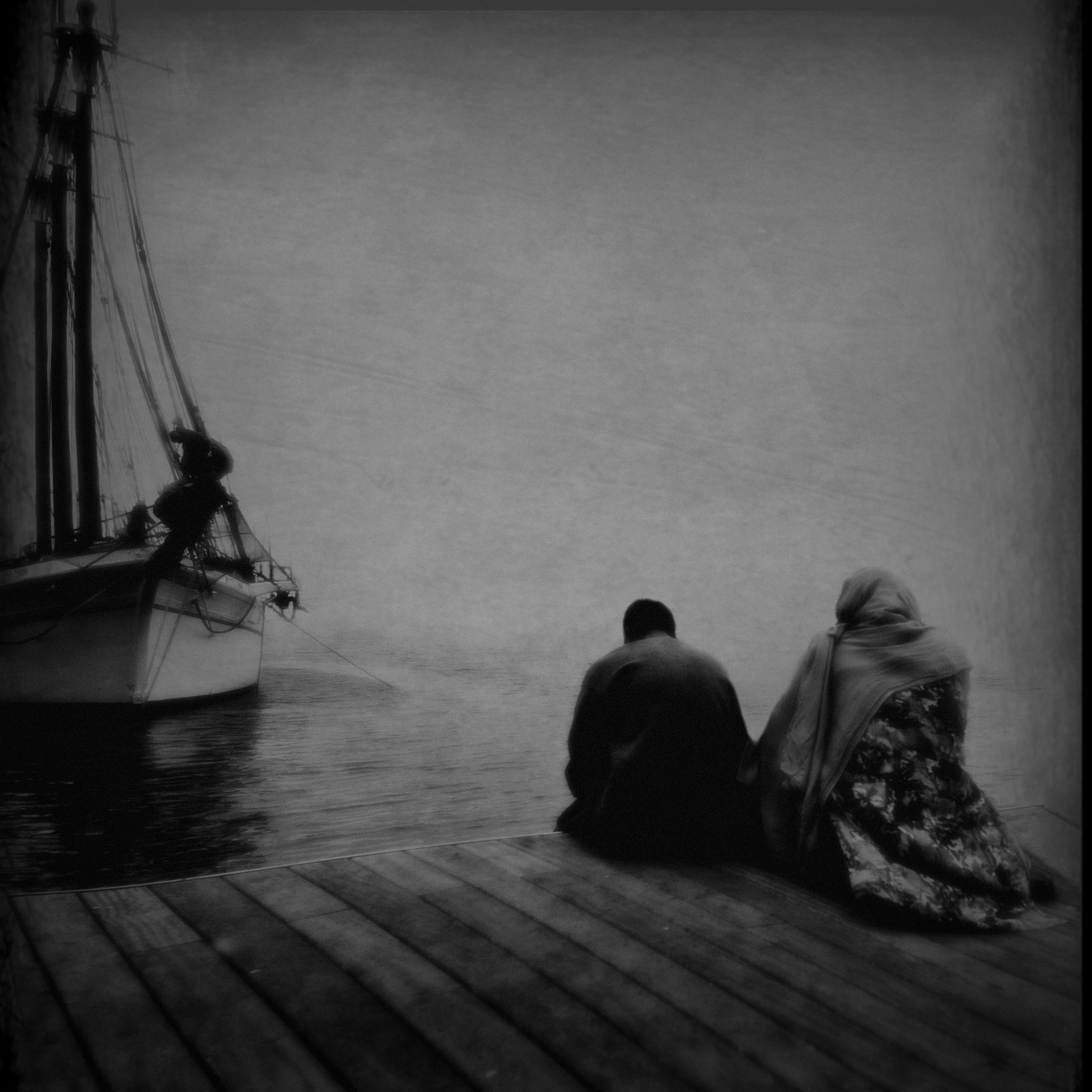 Zwei Menschen sitzen an einem Wasserufer, ein Boot ist im Wasser sichtbar, Stimmung melancholisch im Schwarzweiß-Foto.