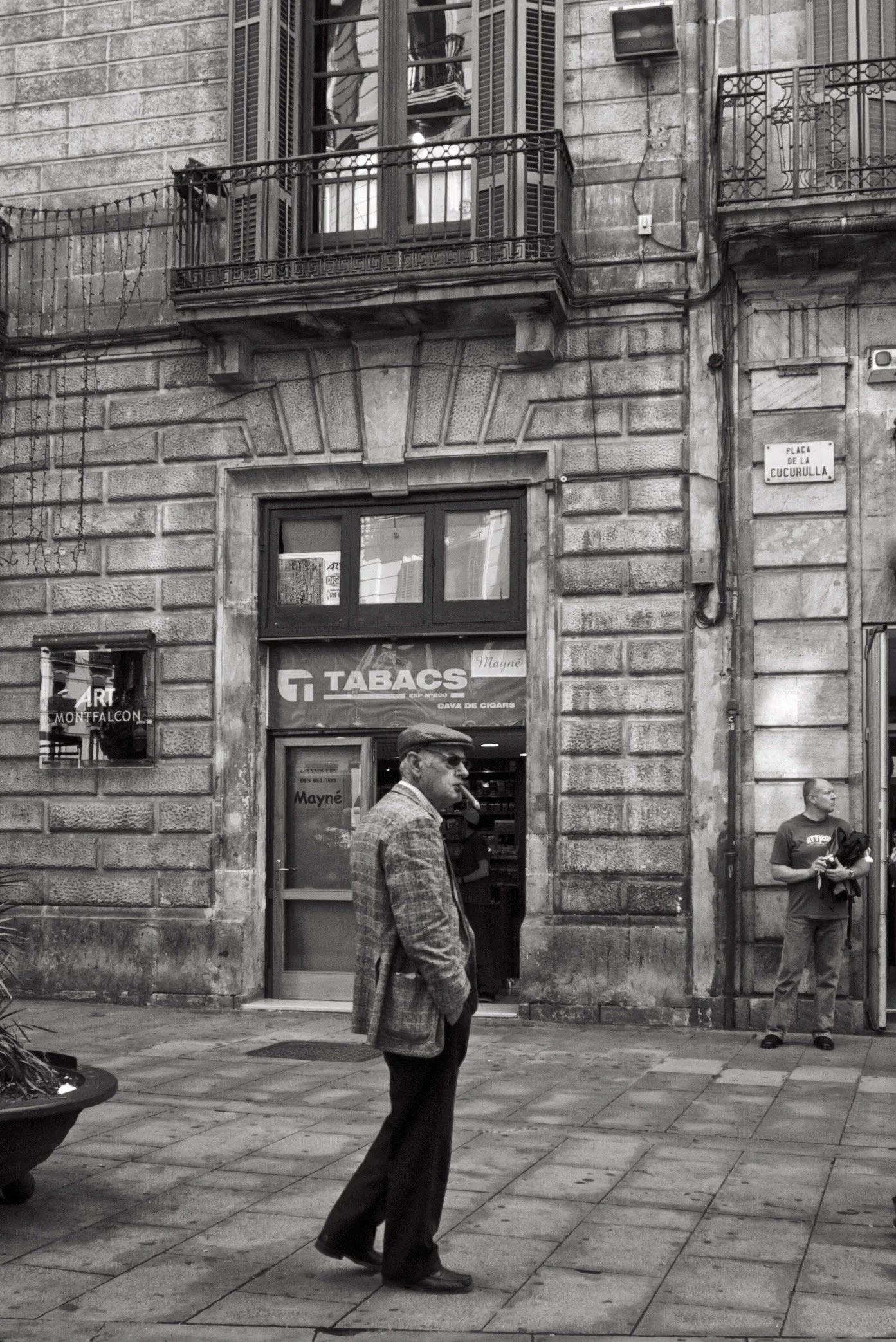 Ein Mann mit einem karierten Jacke und Hut steht auf einer Straße vor einem Geschäft, während im Hintergrund ein zweiter Mann steht, der etwas hält. Das Gebäude hat eine steinerne Fassade mit Balkon und Fenster. Es ist schwarz-weiß fotografiert.