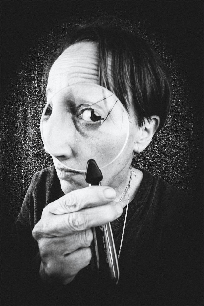 Schwarz-Weiß-Fotografie einer Person, die durch eine Lupe schaut und dabei ein Spiegelbild ihres Gesichts zeigt.