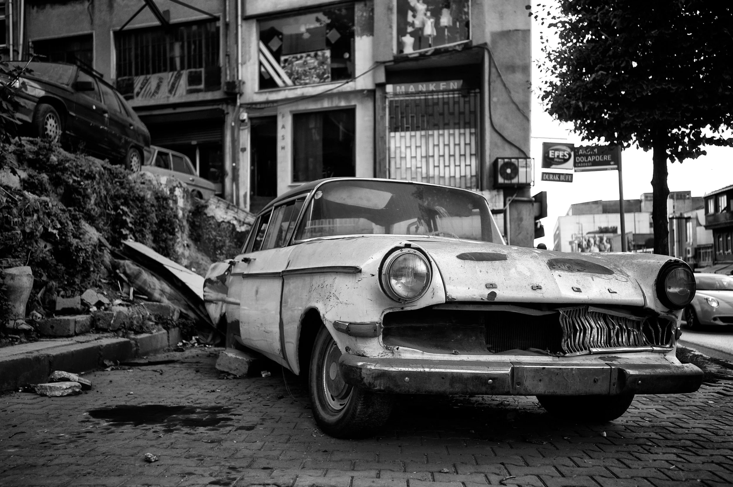 Ein verrosteter, alter Auto steht auf einem Straßenrand vor einem Gebäude, das auf einem Hügel gebaut ist. Der Wagen ist beschädigt und wirkt verlassen.