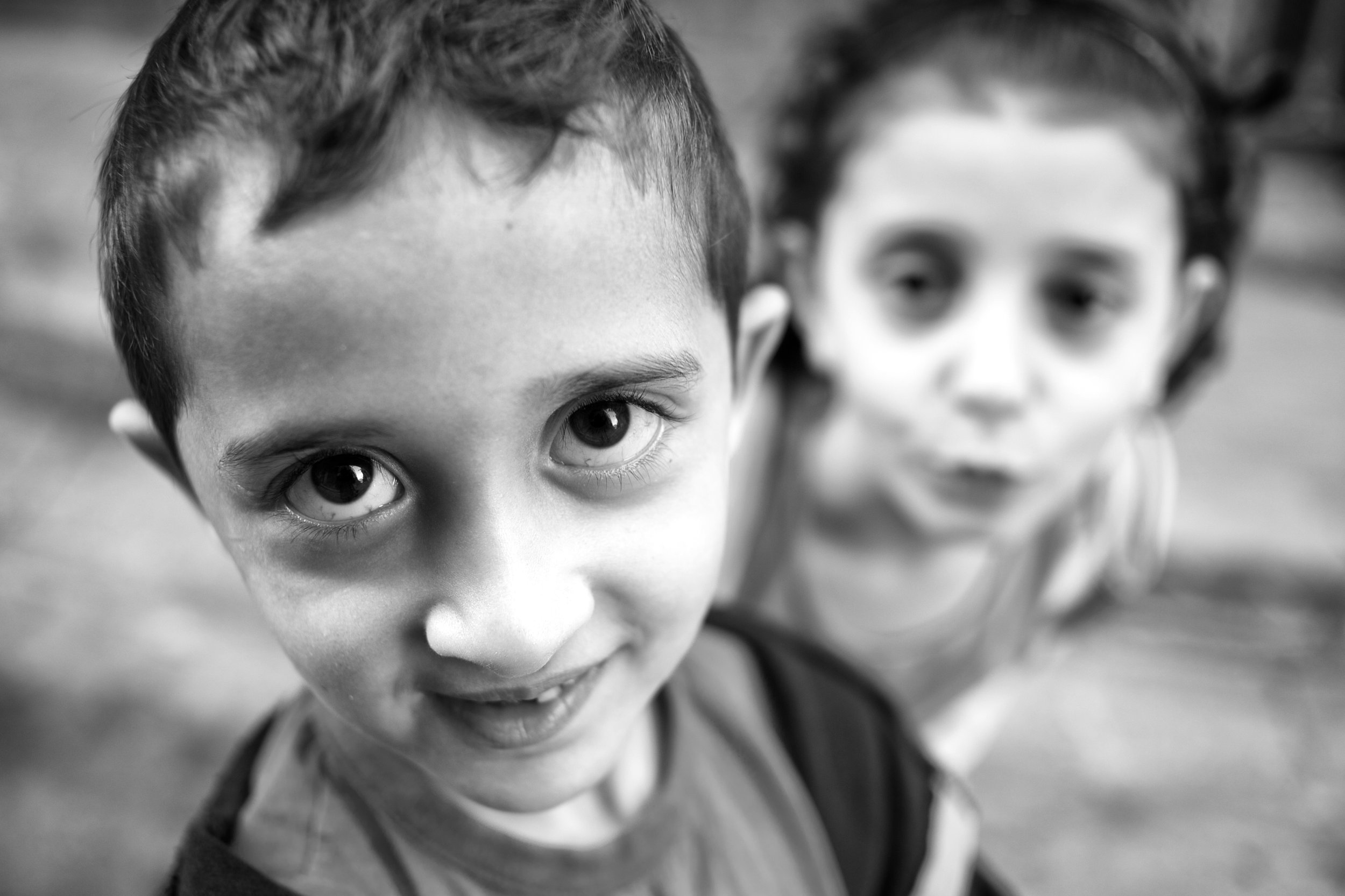 Nahaufnahme von einem Jungen mit großen Augen und einem Lächeln, im Vordergrund, und einem Mädchen im Hintergrund, das ein Gesichtsmasken-Motiv trägt, auf einem Holzboden.