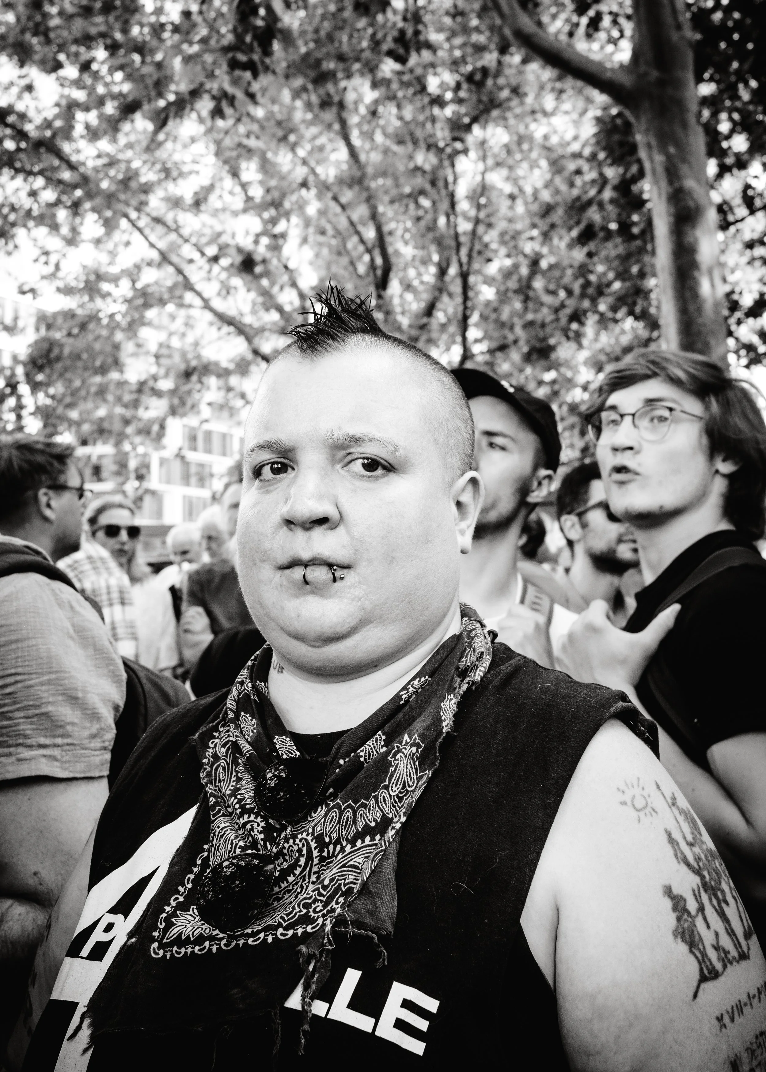 Schwarz-Weiß-Foto einer Person mit Mohawk, Piercings, Tattoos, Sonnenbrille, Bandana und einer Gruppe von Menschen im Hintergrund auf einer Straßendemo in einem Park mit Bäumen.