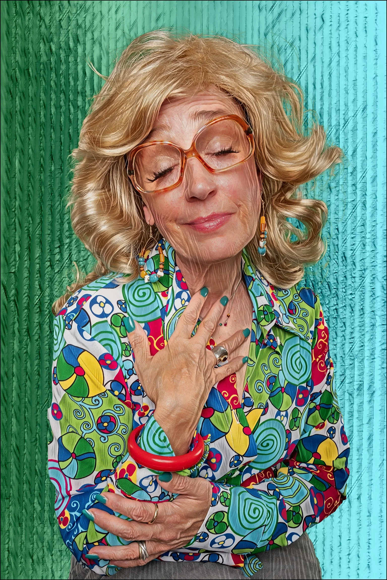 Eine ältere Frau mit blonden, lockigen Haaren trägt bunte Musterkleidung, große Brille, moderne Schmuckstücke, und zeigt eine zufriedene Pose vor einem grünen und blauen Hintergrund.